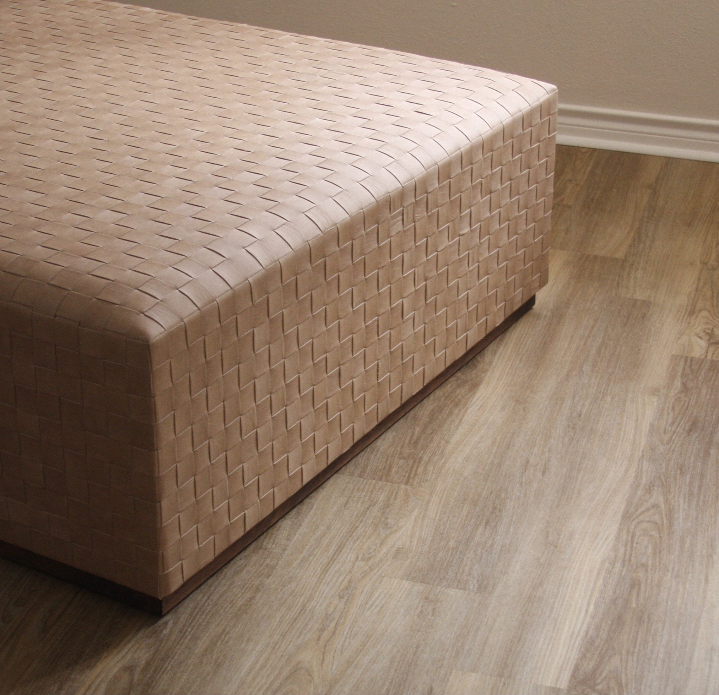 A woven beige rectangular storage ottoman on wooden flooring next to a beige wall with white baseboard.