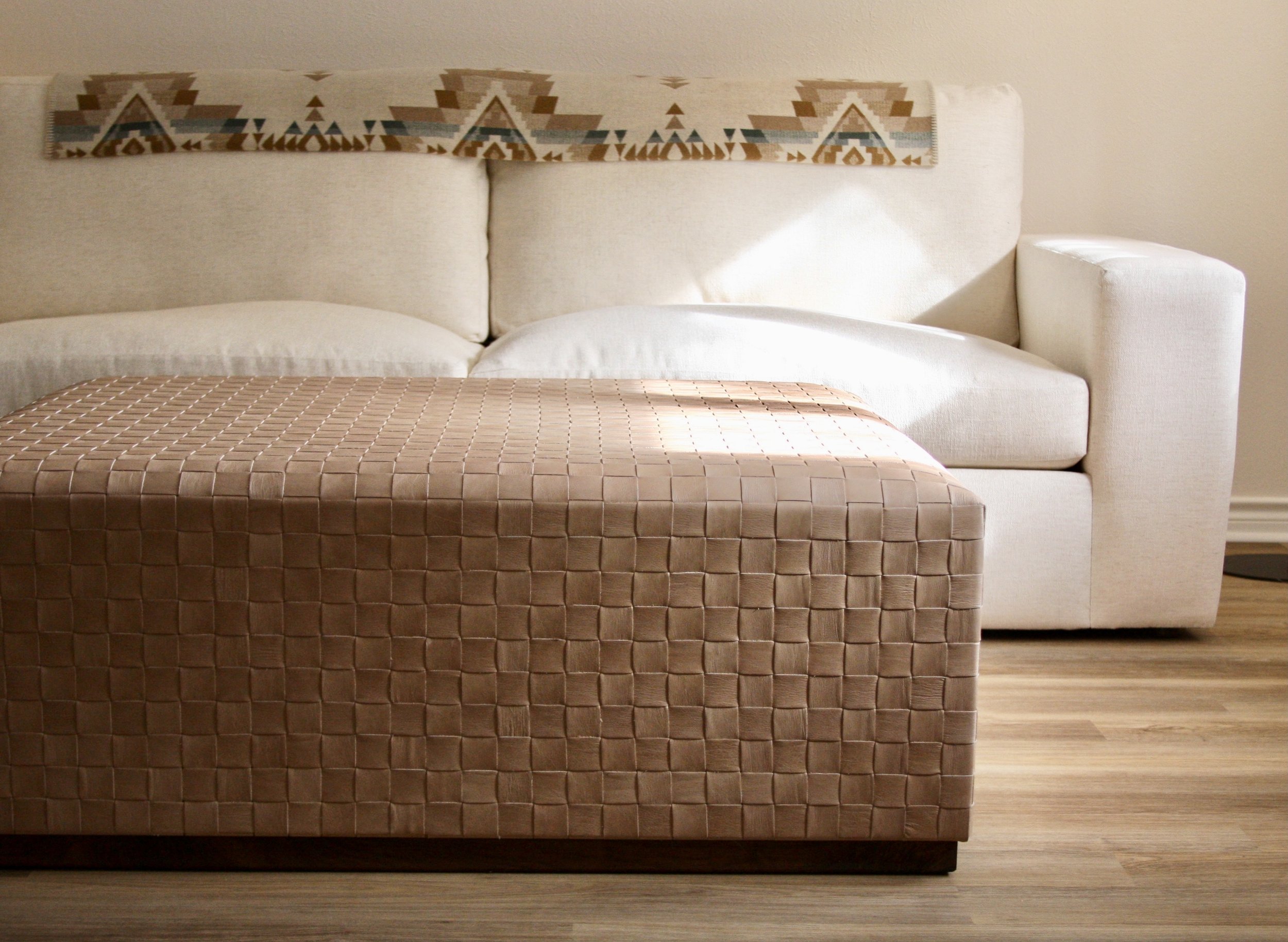 A beige sofa with a Southwestern-style patterned backrest and a woven beige ottoman in front of it, on a wooden floor.