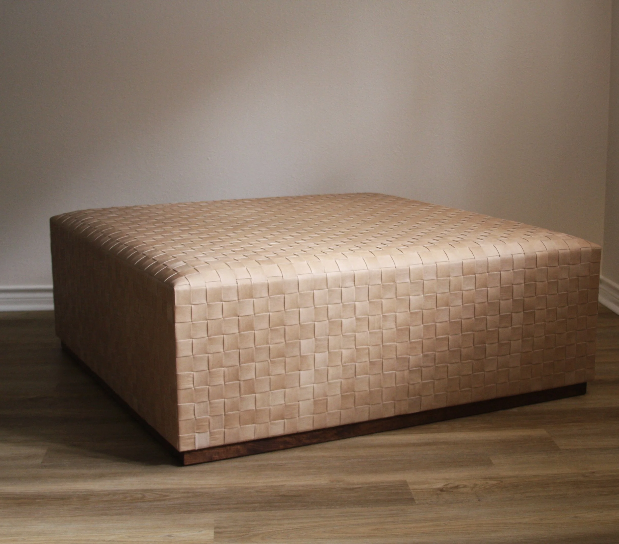 A beige woven textile ottoman with a wooden base against a plain wall.