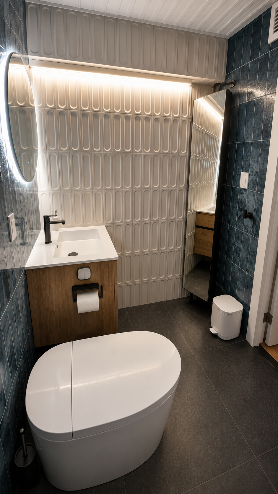 Modern bathroom with a white, oval freestanding toilet, dark tiled floor, wooden vanity with a white sink, round mirror with LED light, patterned wall tiles, and a small trash bin.