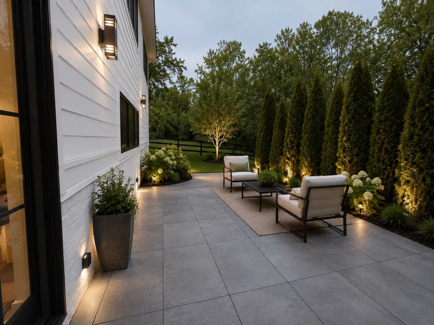 Outdoor patio with two cushioned chairs, a small black table with a potted green plant, a beige rug, potted plants along the wall, a tree with light-colored leaves, and a row of tall evergreen trees in the background illuminated by ground lights at d