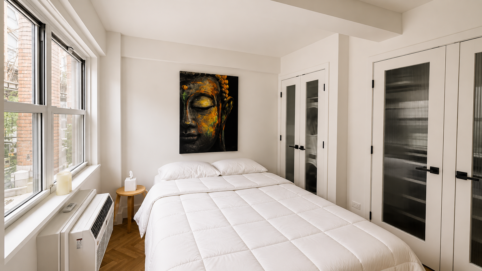 A minimalist bedroom with a large window, a white bed with pillows and a quilt, wall art of a Buddha face, and built-in closets with black handles.