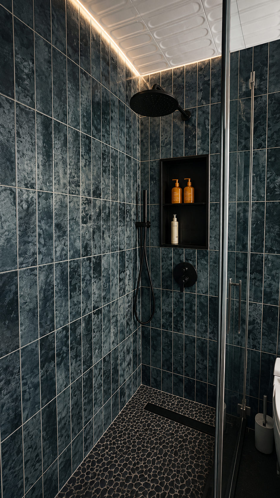 Modern shower with dark green marbled tiles, black ceiling shower head, black handheld shower, built-in black shelf with two bottles of yellow soap and one white bottle, black pineapple-shaped drain cover on textured pebble floor, and a glass partition with a metal handle.