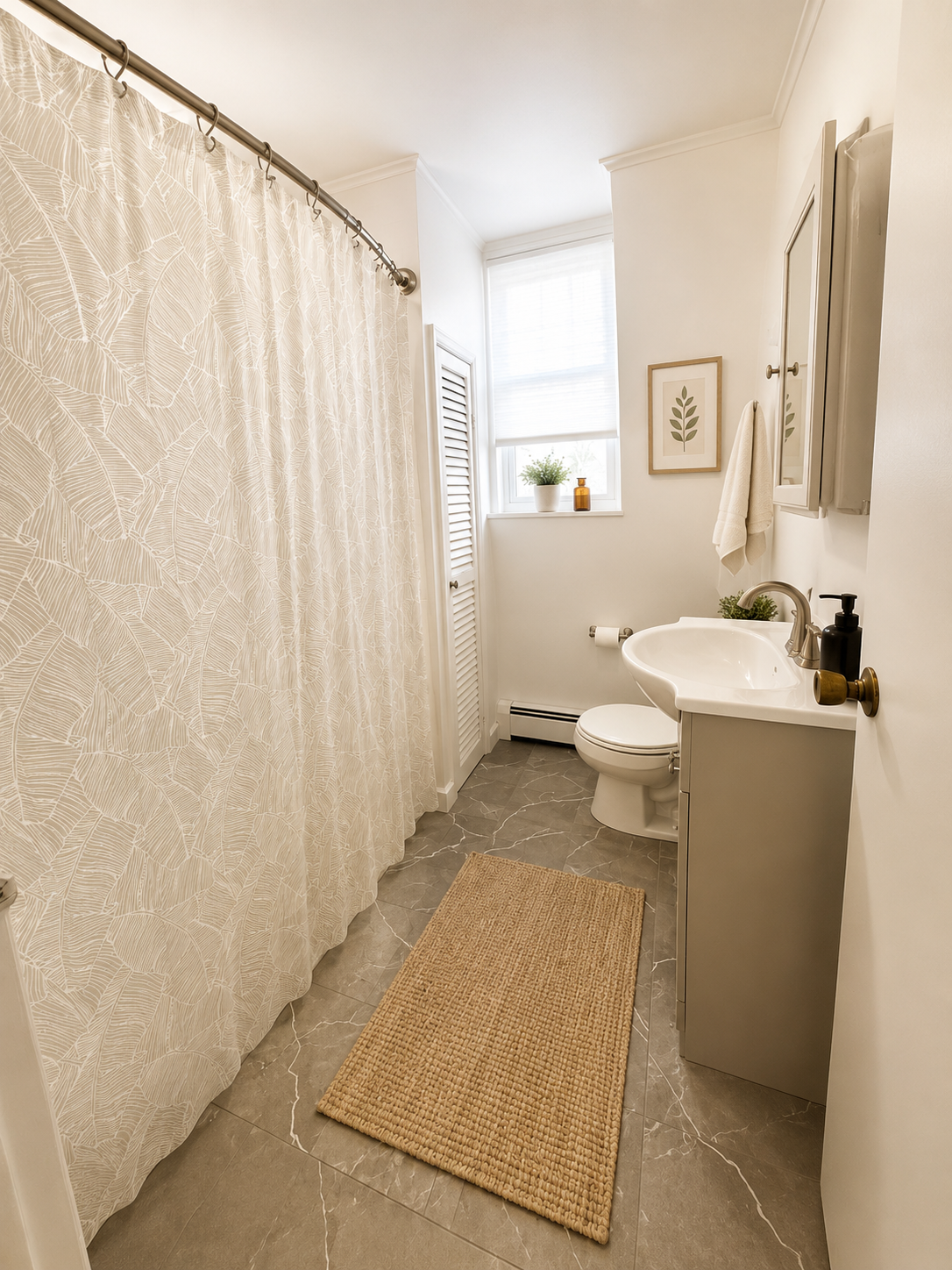 Small bathroom with beige and white decor, shower with leaf-patterned curtain, toilet, small sink, window with blinds, and wall decorations.