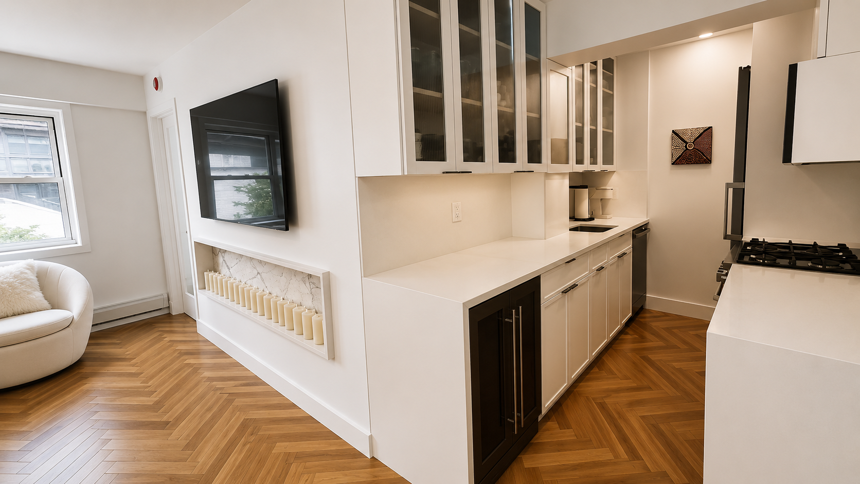 Modern kitchen with white cabinets, black bottom cabinet, and wood herringbone flooring. The counter has a small sink and a coffee maker. There's a wall art piece and a view of the living area with a white couch and a TV on the wall.