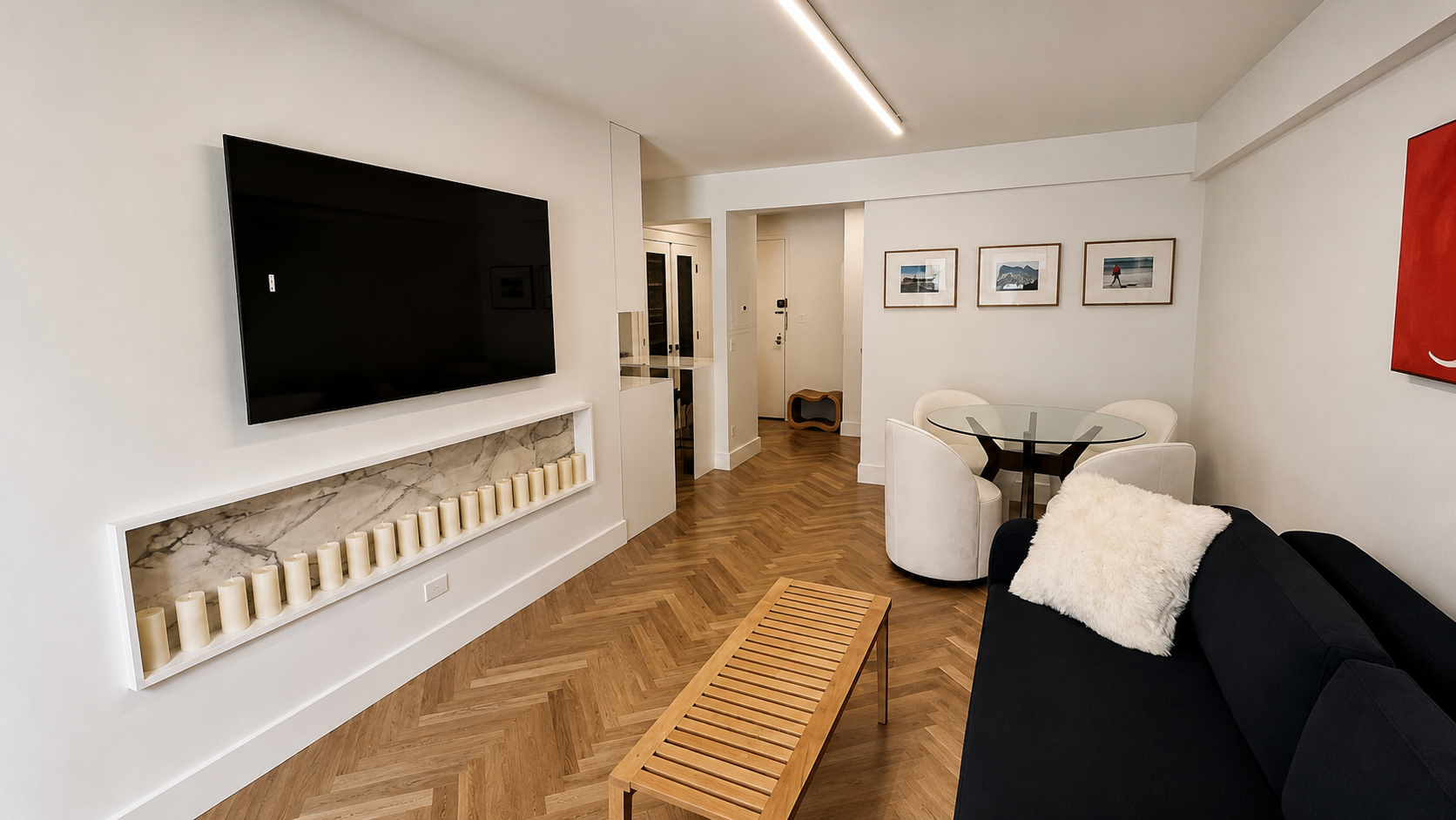 Living room with a large flat-screen TV on the white wall, a wooden bench, a black sofa with a white fluffy pillow, a white round dining table with three white chairs, wall art with four framed pictures, and candles on a marble fireplace mantel.