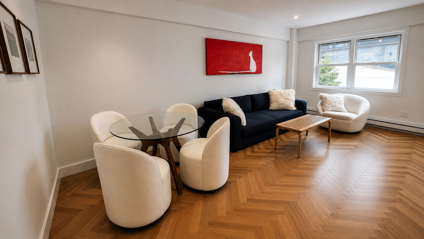 A living room with a black sofa, a white armchair, and a round glass dining table with four white chairs. There's a wooden coffee table in front of the sofa, a window allowing natural light, and wall art including a red painting and three smaller bla