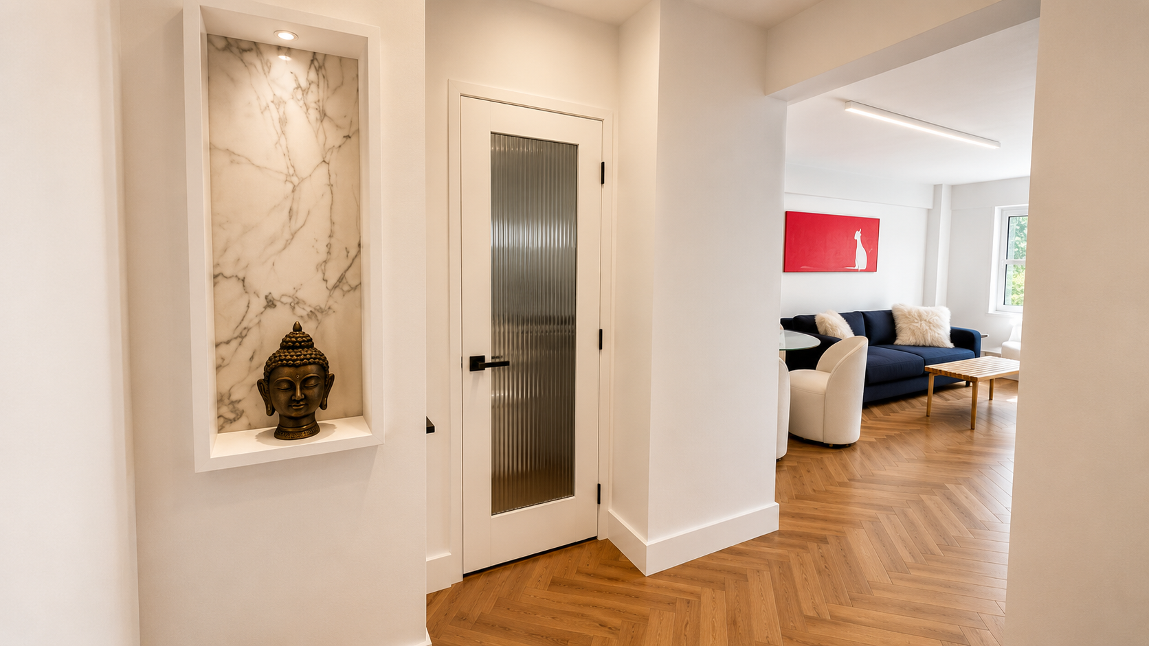 Interior of a modern apartment with a hallway, white walls, and a decorative alcove with a Buddha head statue. The hallway leads to a living room with a navy sofa, white fluffy pillows, a black armchair, a wooden coffee table, and a red artwork on th