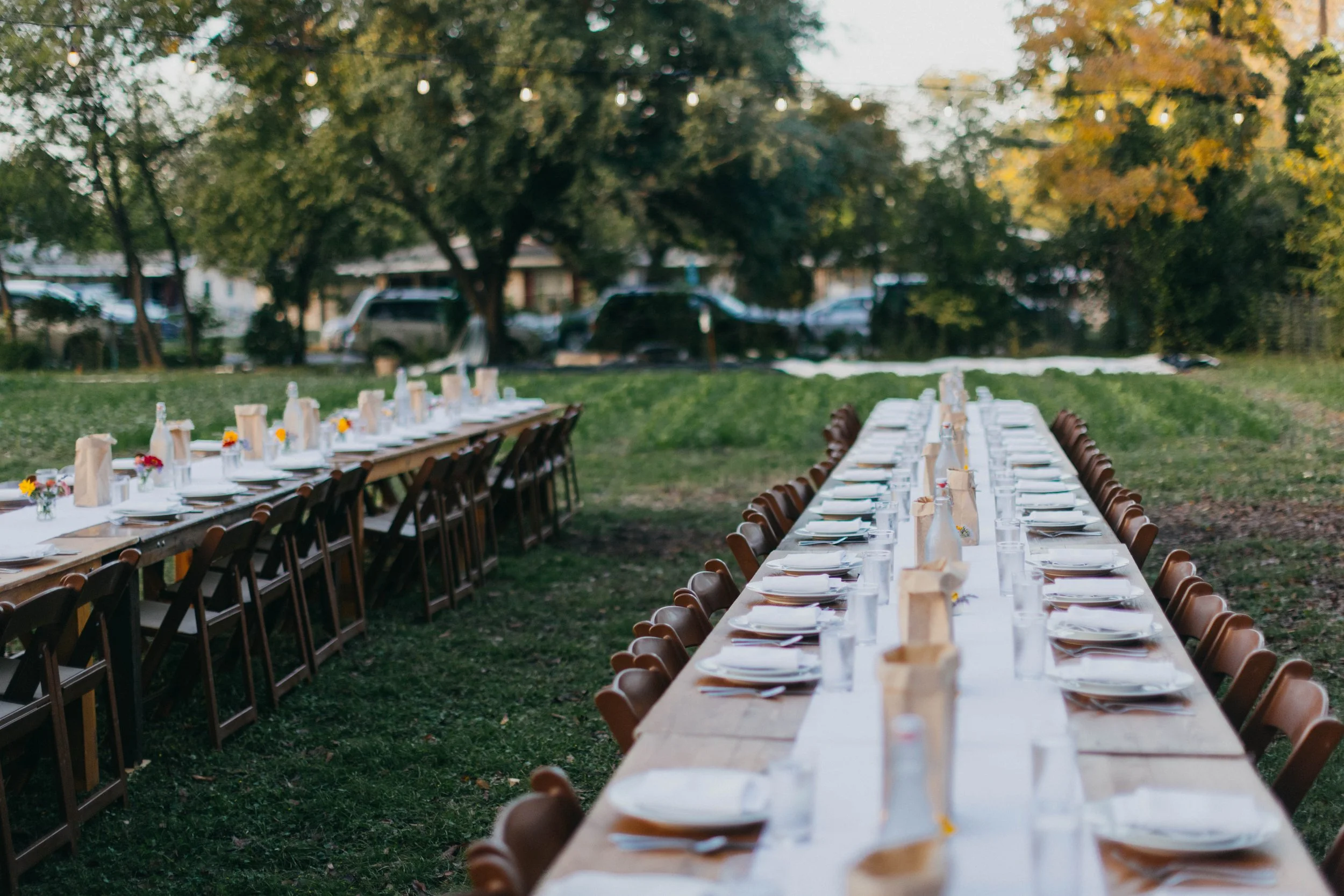 Long outdoor dining tables set with plates, glasses, napkins, and small flower arrangements, surrounded by wooden chairs, in a grassy area with trees and parked cars in the background.