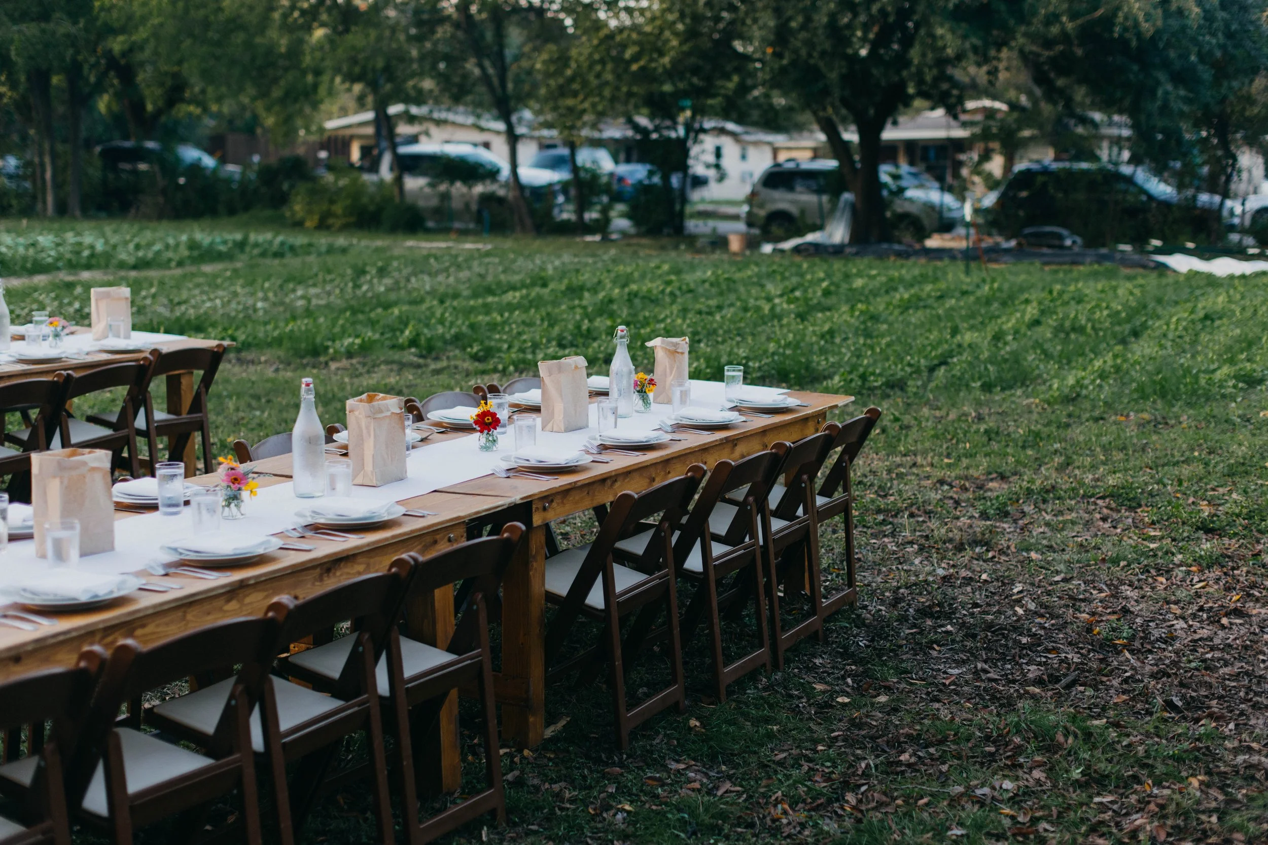 An outdoor dining setup with a long wooden table, set with plates, glasses, napkins, small flower arrangements, and water bottles on a grassy area surrounded by trees.