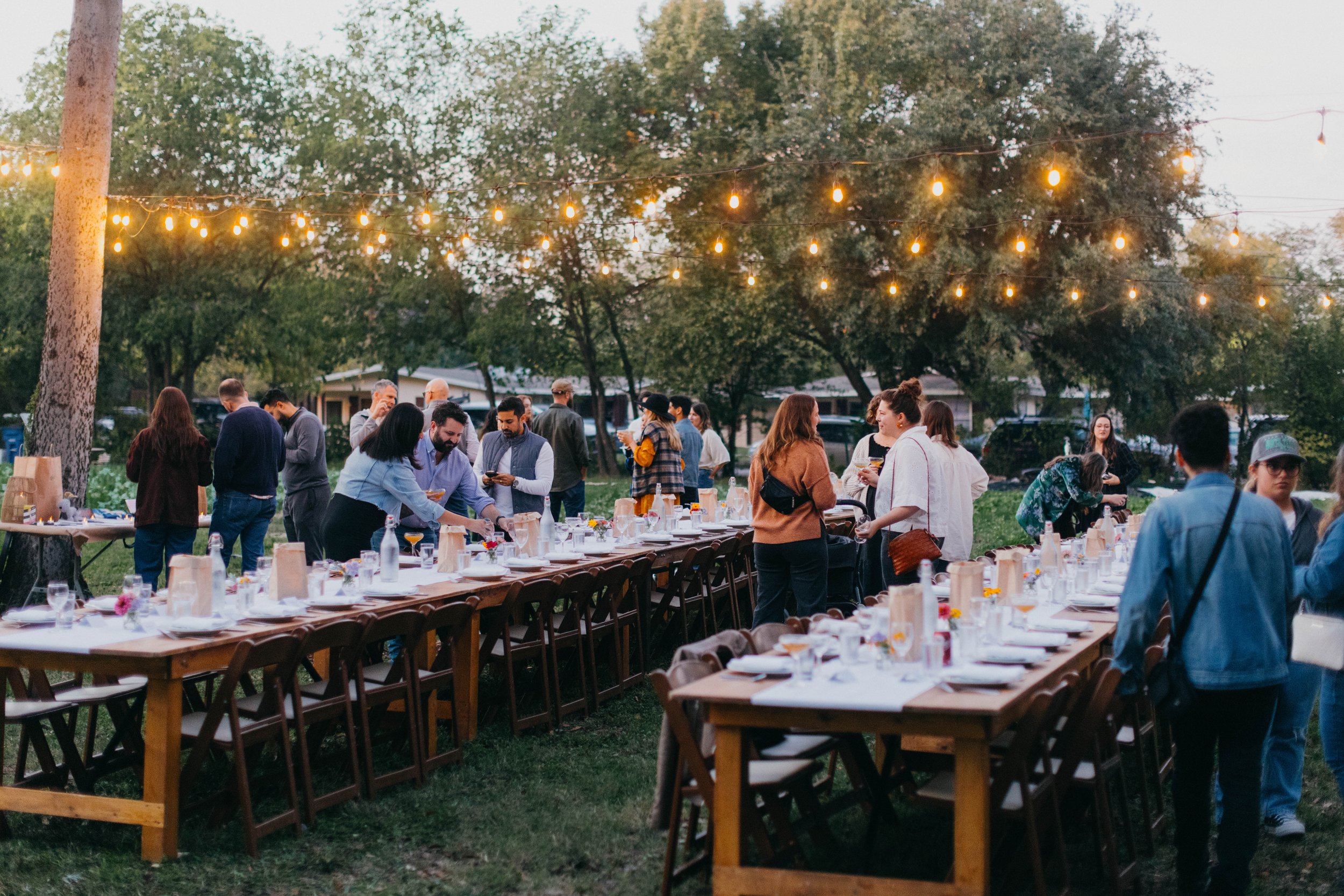 Outdoor gathering with long dining tables set with white tablecloths, glasses, and centerpieces, under string lights in a green park area, with people mingling and socializing in the evening.