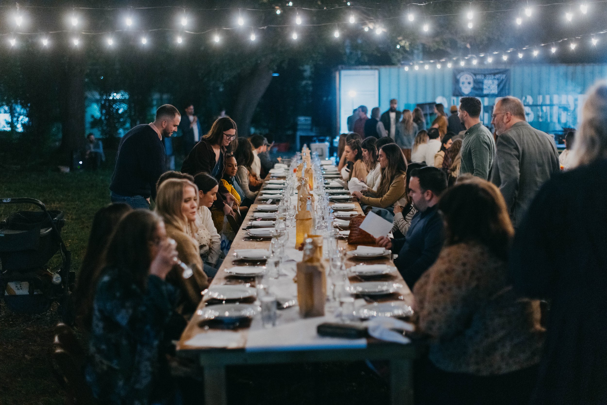 Outdoor evening event with a long table of people dining under string lights.
