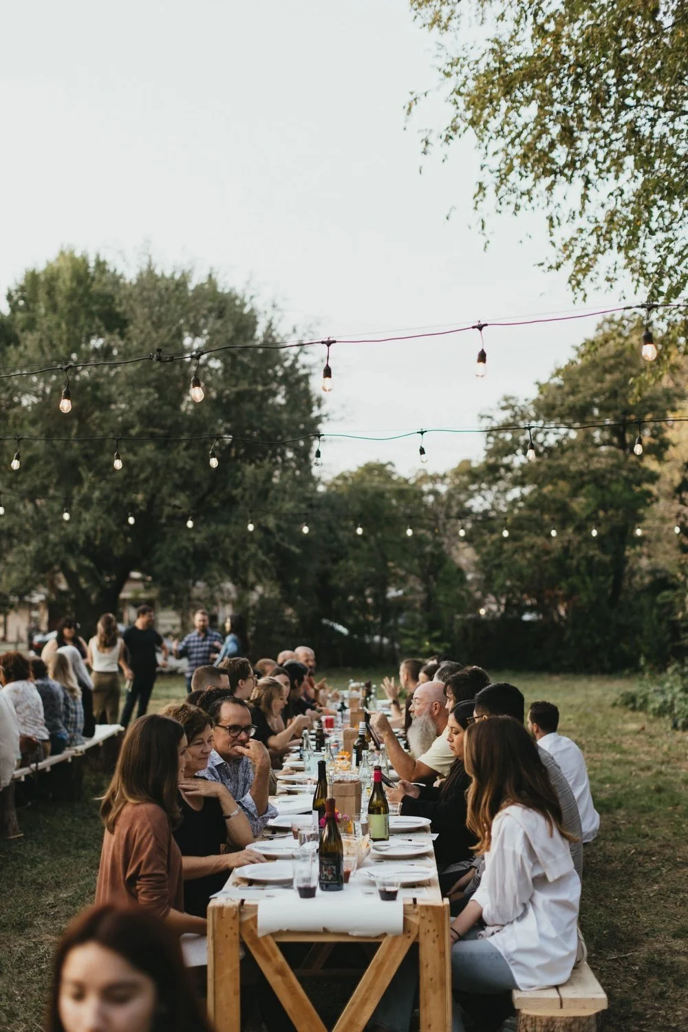 A large outdoor gathering with people sitting at a long table, many engaged in conversation, with string lights hanging above, surrounded by trees.
