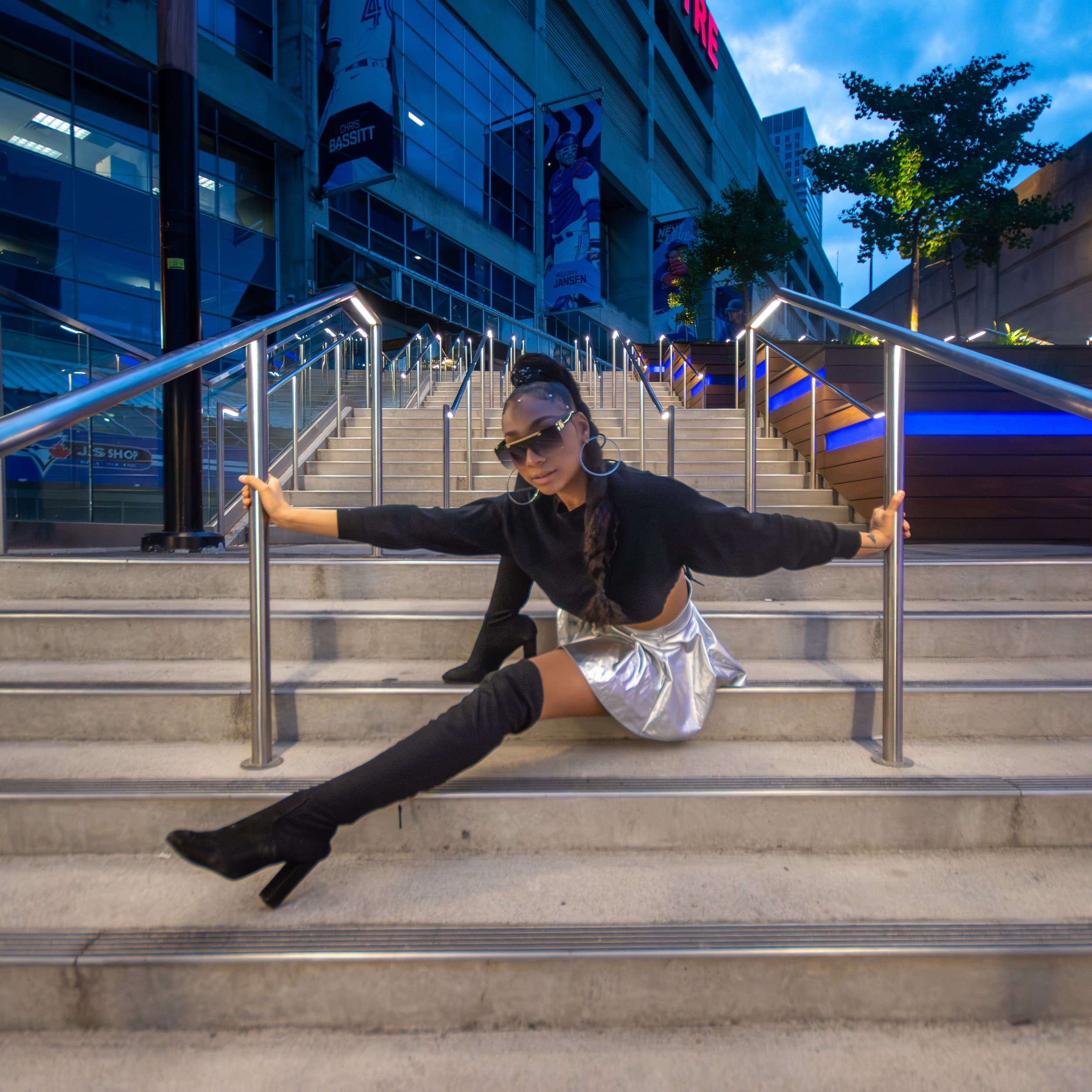 A woman with dark hair in a ponytail and wearing sunglasses poses on outdoor stairs with glass railings, in front of a modern building with large windows and banners of hockey players.
