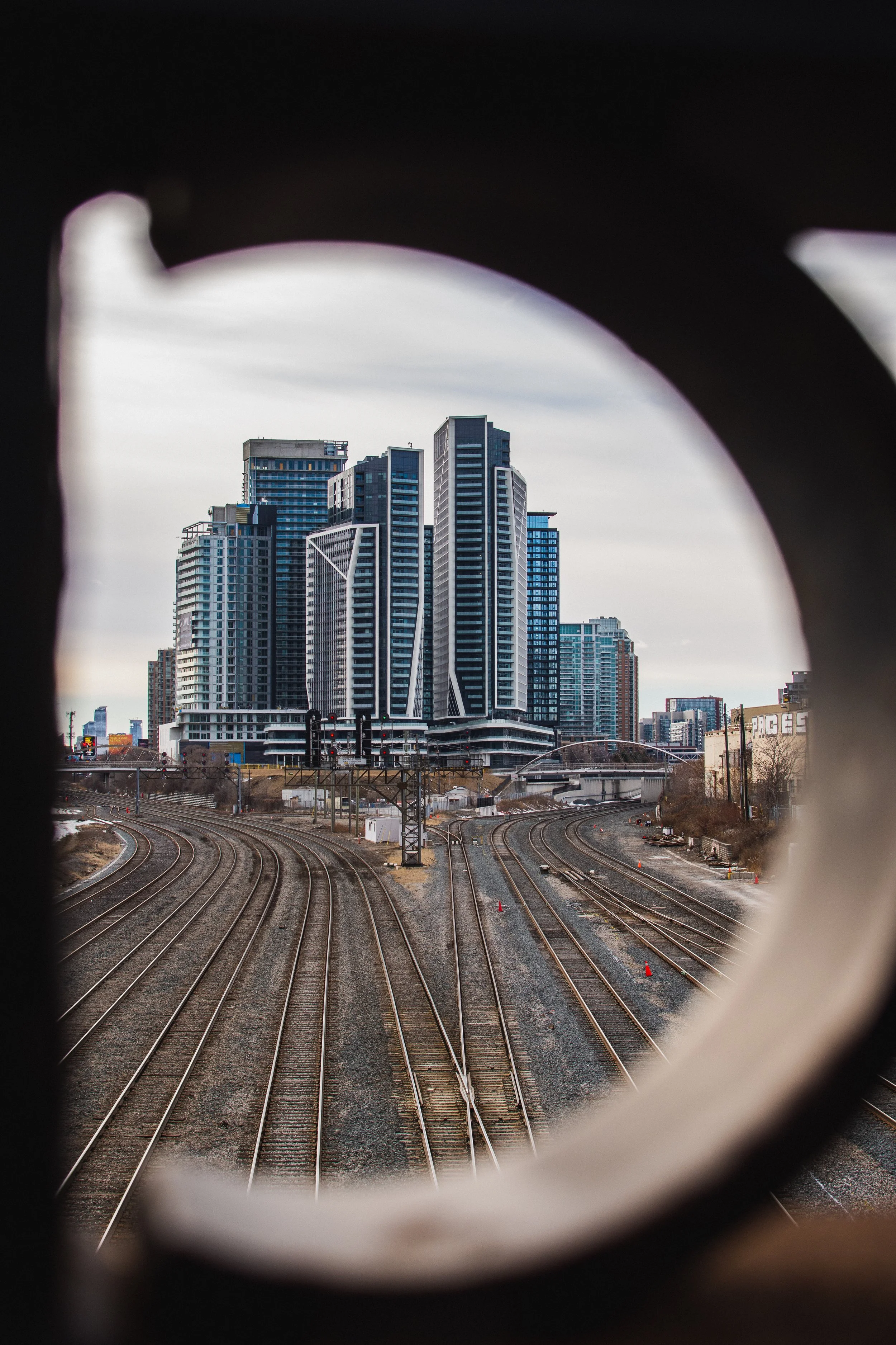 City skyline with tall modern buildings viewed through a circular opening in a barrier, with railway tracks in the foreground.