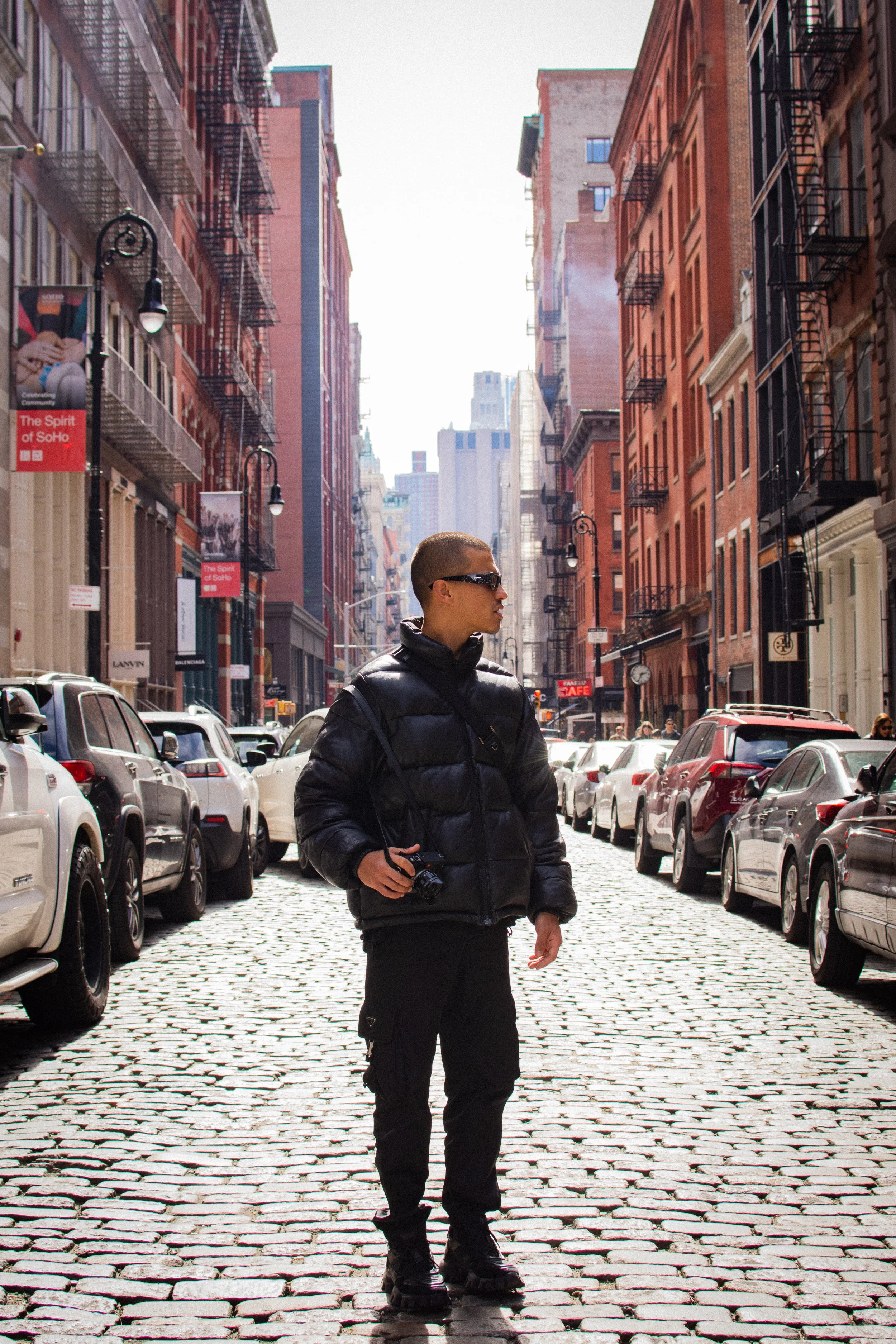 A man in black sunglasses and a puffy black jacket stands on a cobblestone street in a city, holding a camera, with parked cars on both sides and tall brick buildings with fire escapes.