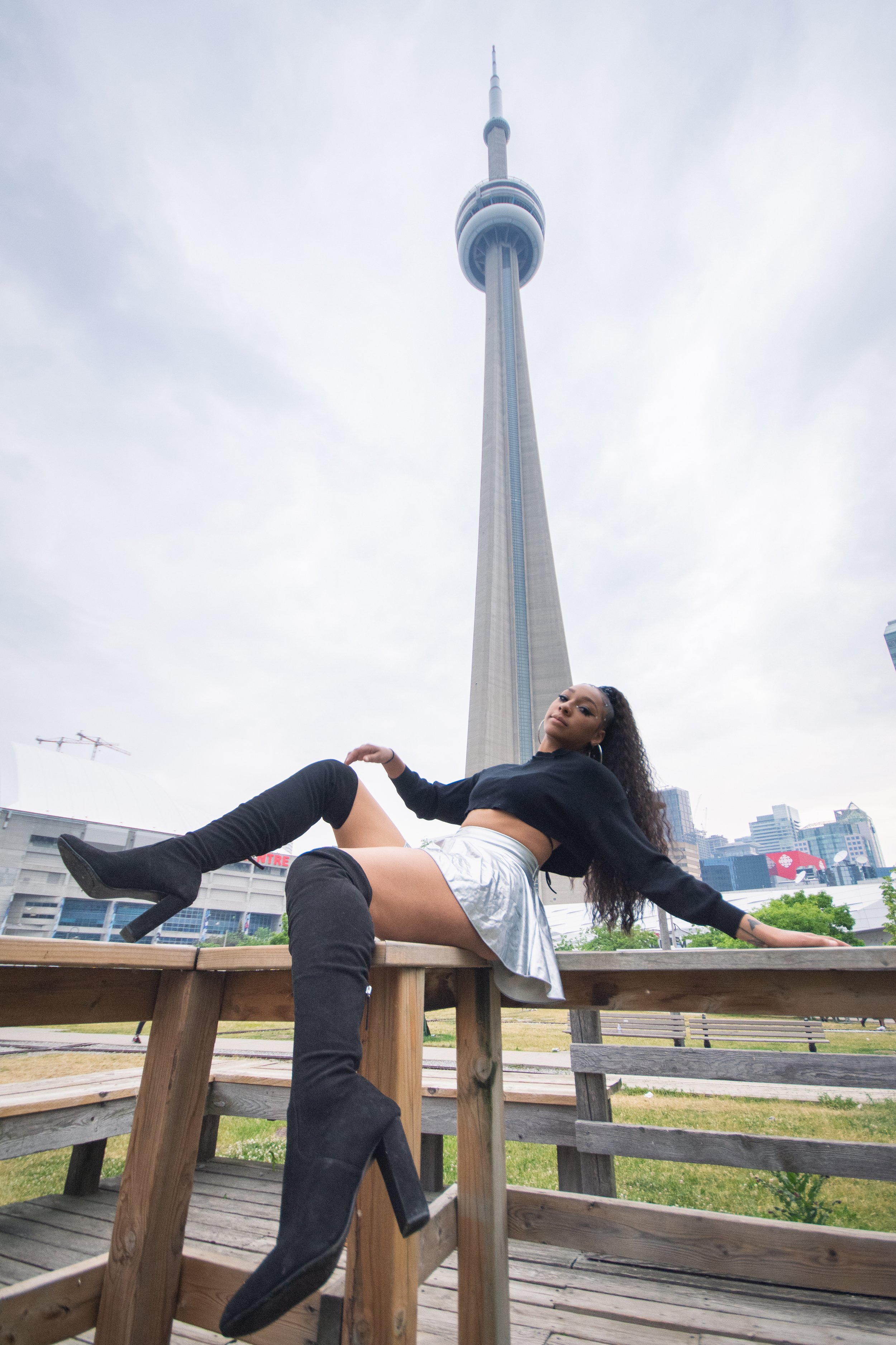 A woman with curly hair wearing a black crop top, metallic shorts, and black thigh-high boots sitting on a wooden outdoor table in front of the CN Tower in Toronto, Canada.