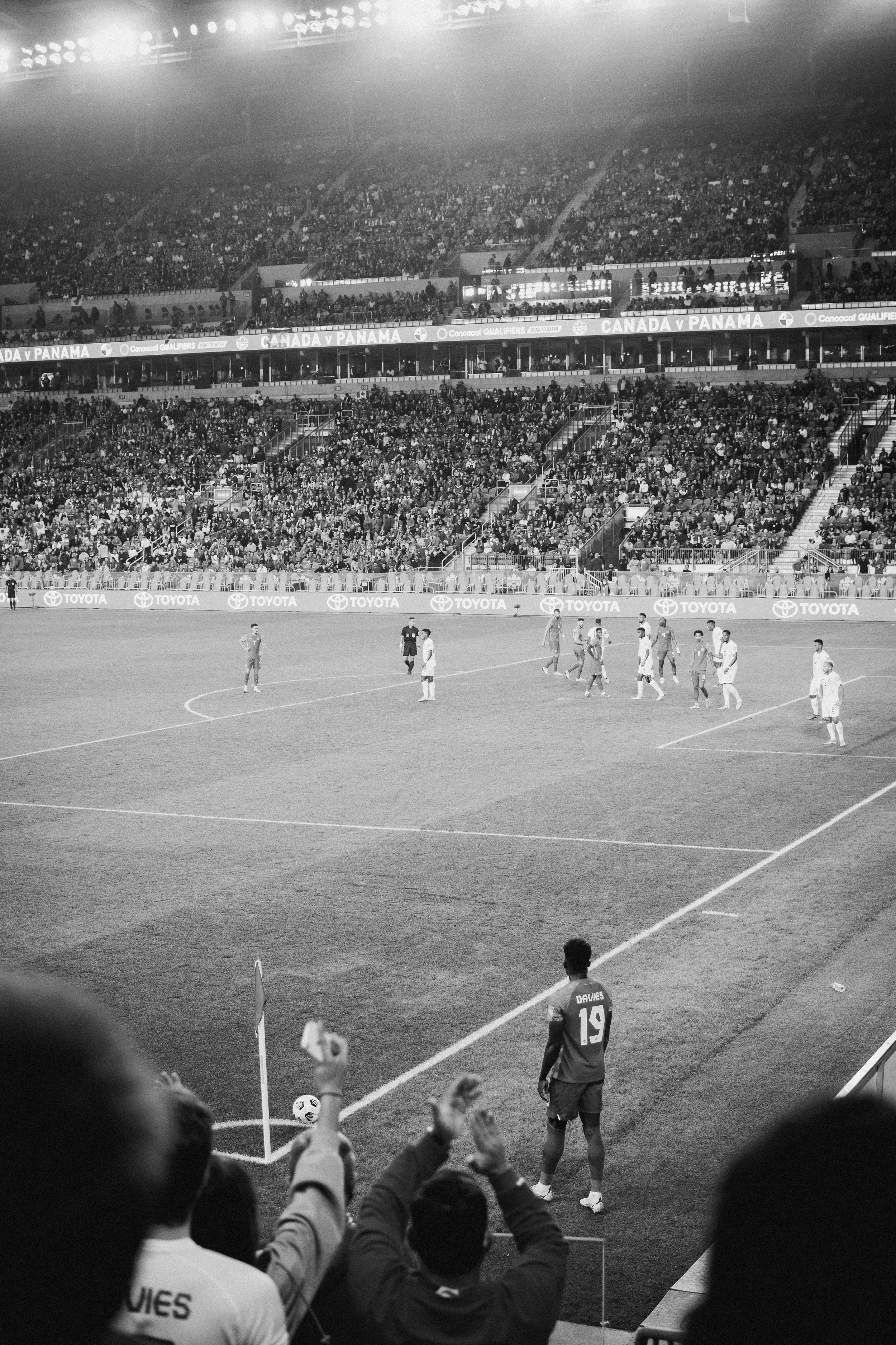 A soccer match is taking place in a stadium filled with spectators. Players are on the field, and fans are waving their arms and cheering. Some players stand near the corner flag, preparing for a game restart.