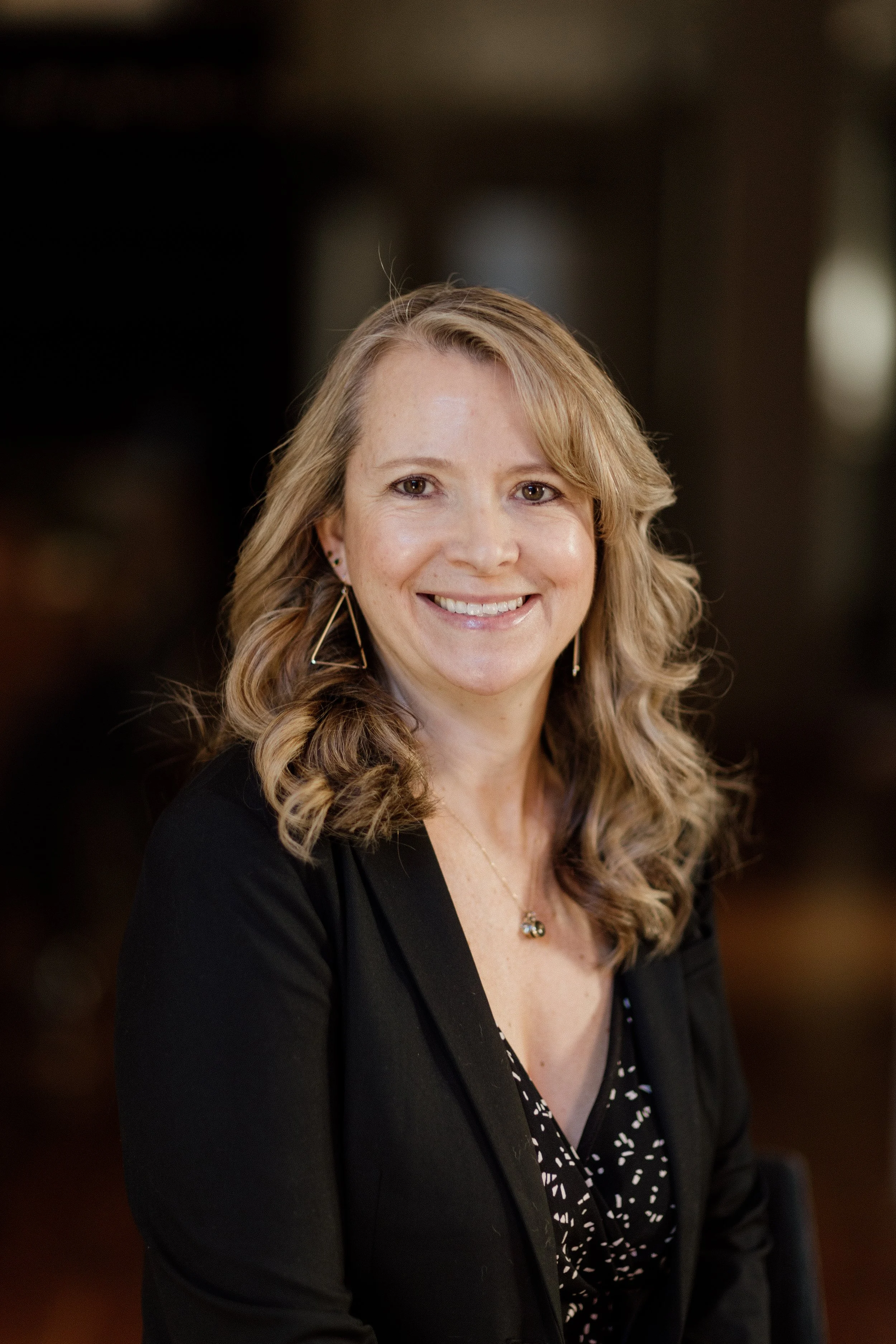 A woman with wavy blonde hair, smiling, wearing earrings, a necklace, a black blazer, and a patterned black and white shirt, in an indoor setting.