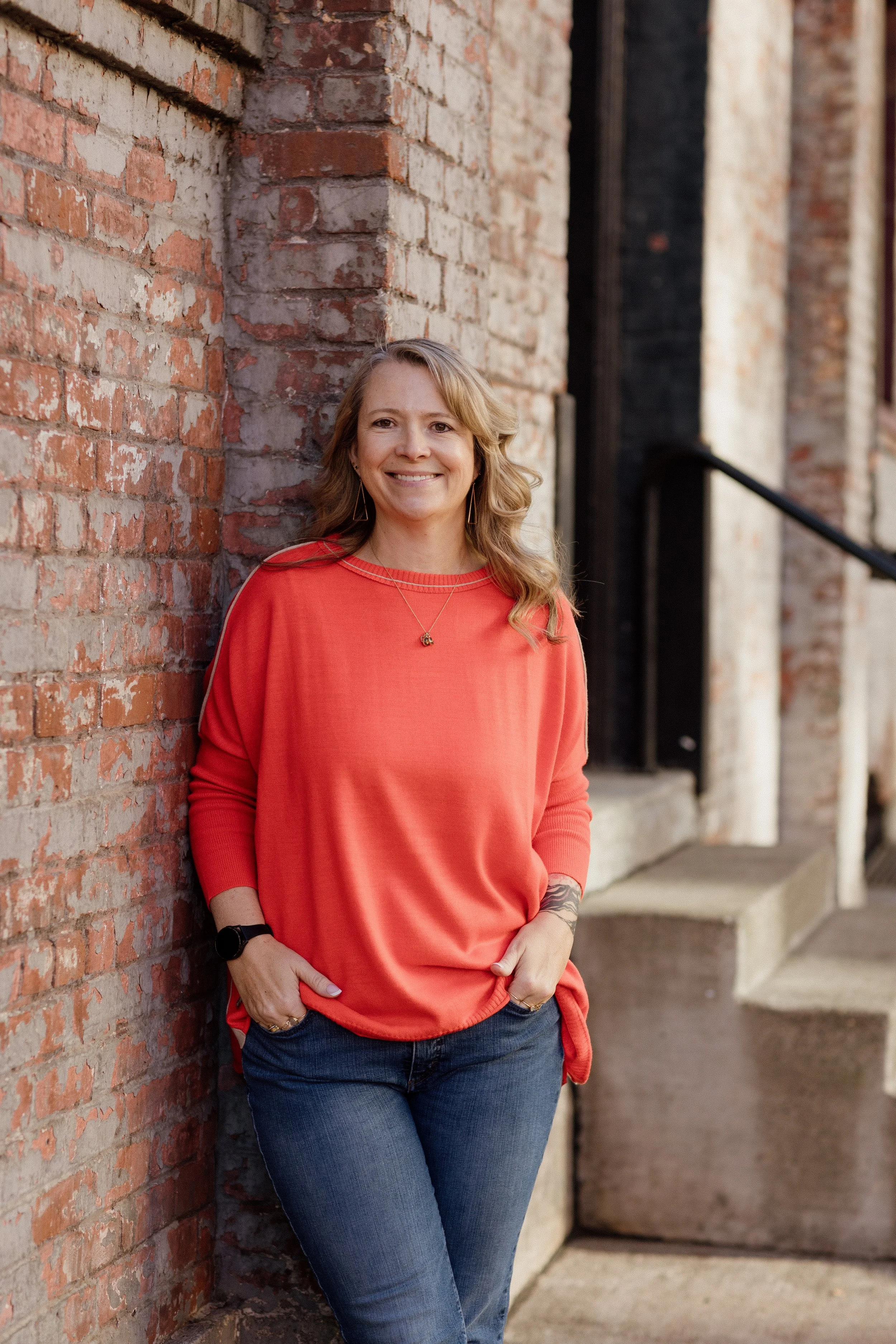 A woman with wavy blonde hair wearing a red long-sleeve shirt and blue jeans, smiling, and standing beside a brick wall with a black handrail in the background.