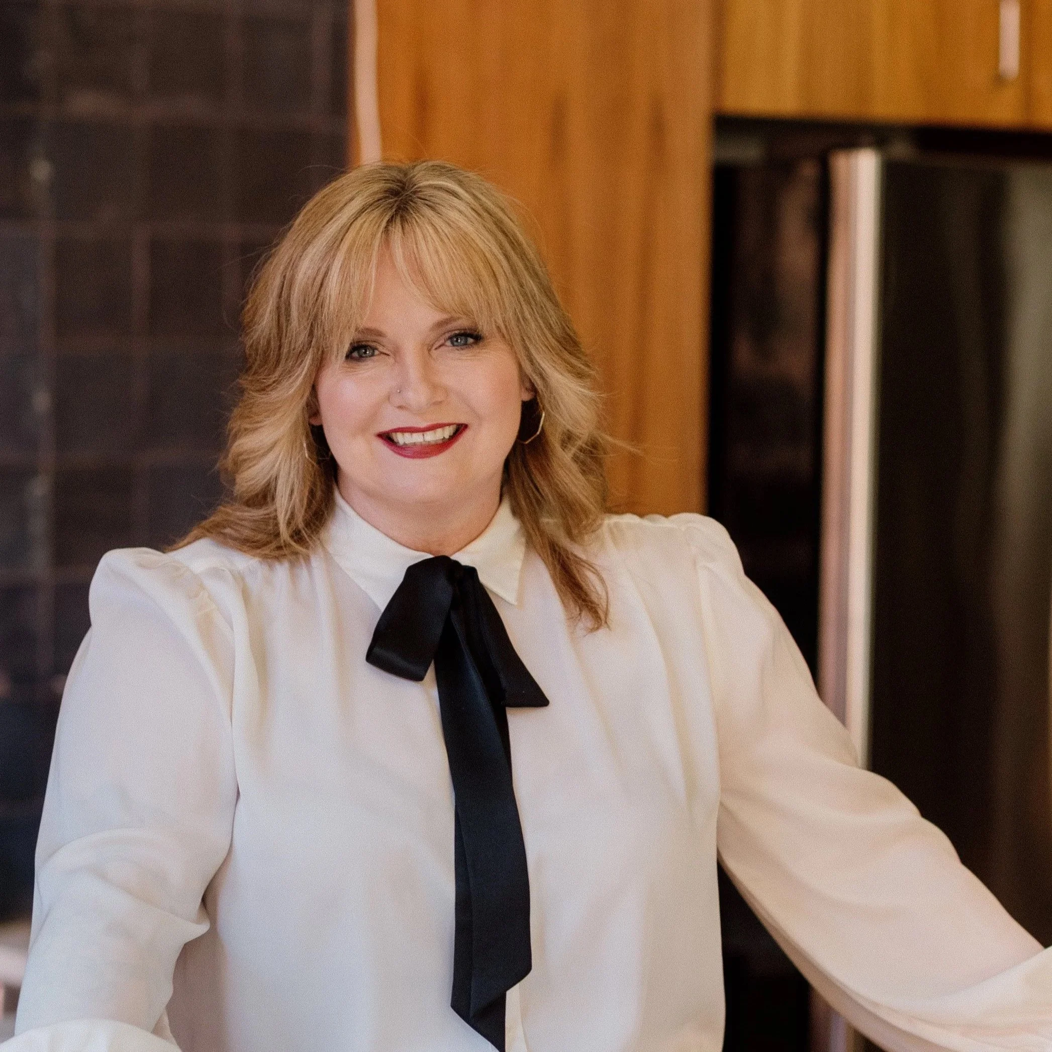 A woman with blonde hair, red lipstick, and hoop earrings, wearing a white shirt with a black bow tie, smiling in a kitchen with wooden cabinets and dark tiled backsplash.