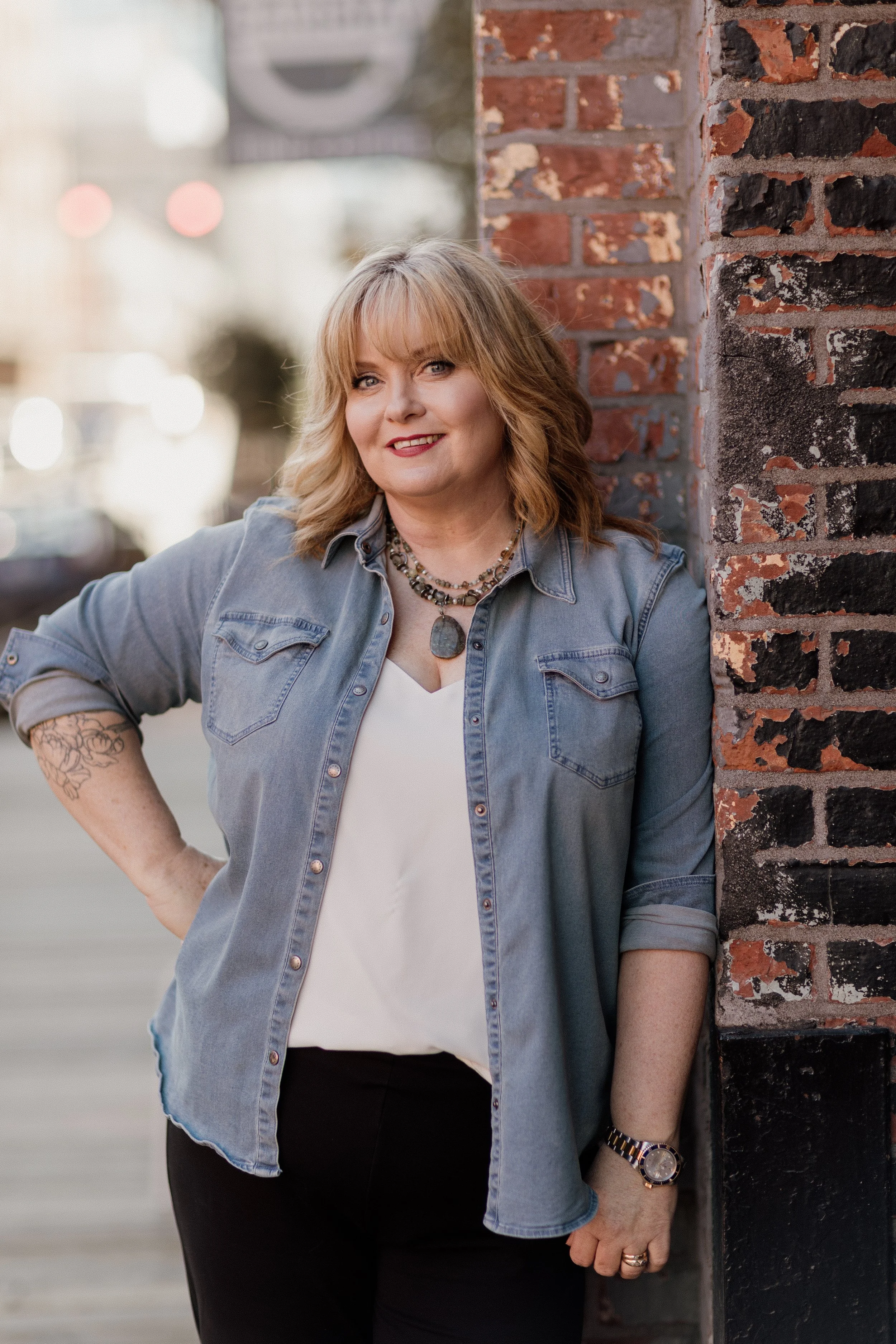 A woman with shoulder-length blonde hair, blue eyes, and a tattoo on her right forearm, wearing a denim shirt, black pants, layered necklaces, a watch, and rings, leaning against a brick wall outdoors, smiling at the camera.