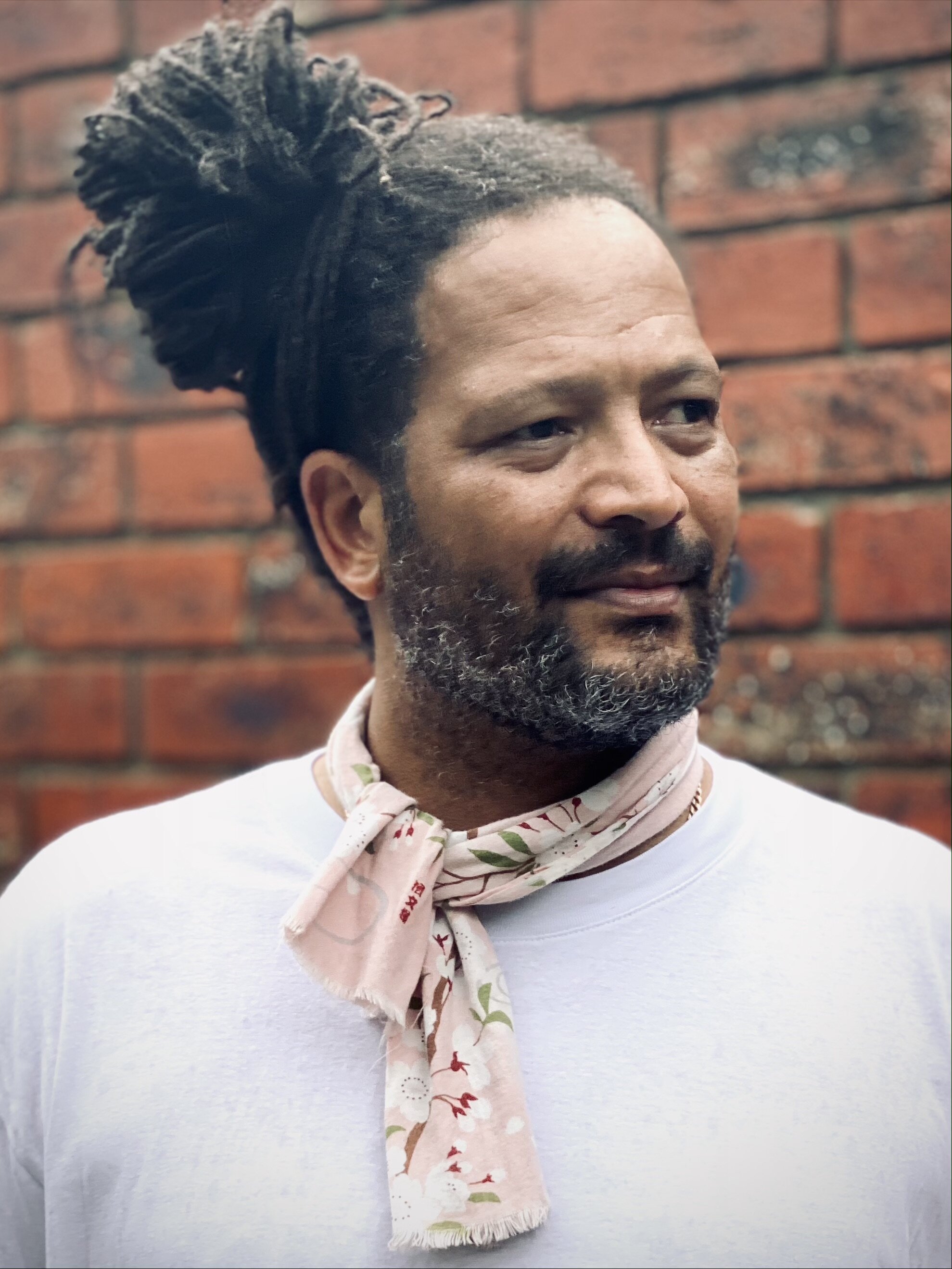 Image of Christian Biko standing in front of a brick wall wearing a white shirt and Japanese style scarf called a Tenugui.