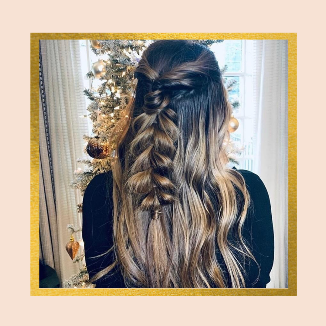 Back view of a person with long, wavy brown hair styled in a half-up hairstyle with a braid, standing in front of a decorated Christmas tree indoors.