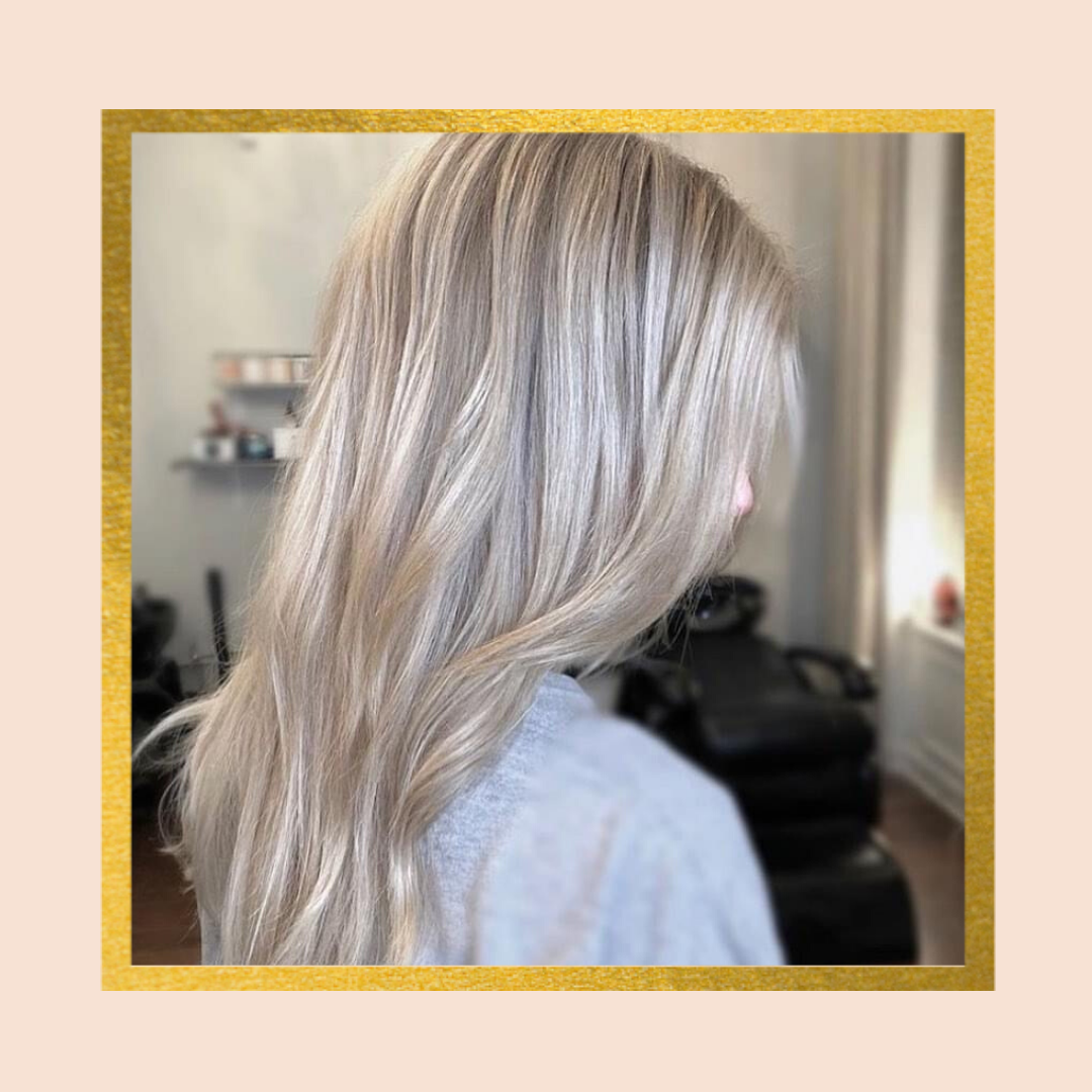 A person with long, ash blonde hair styled in loose waves, set in a soft-focus, salon-like environment.
