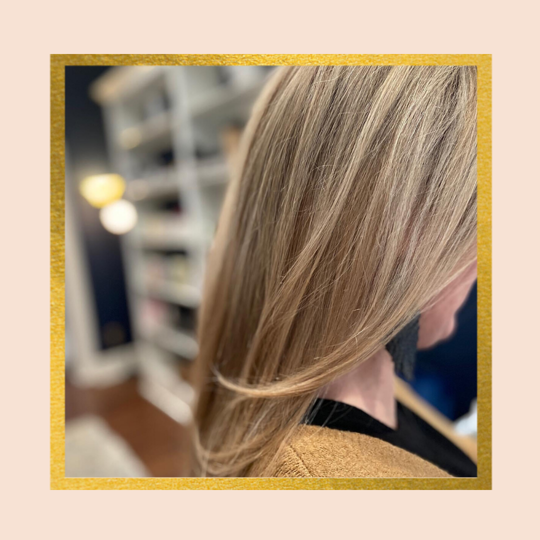 Close-up of a woman with long blonde hair, styled straight, with a blurred background of indoor shelves and a lamp. Framed with a gold border on a beige background.