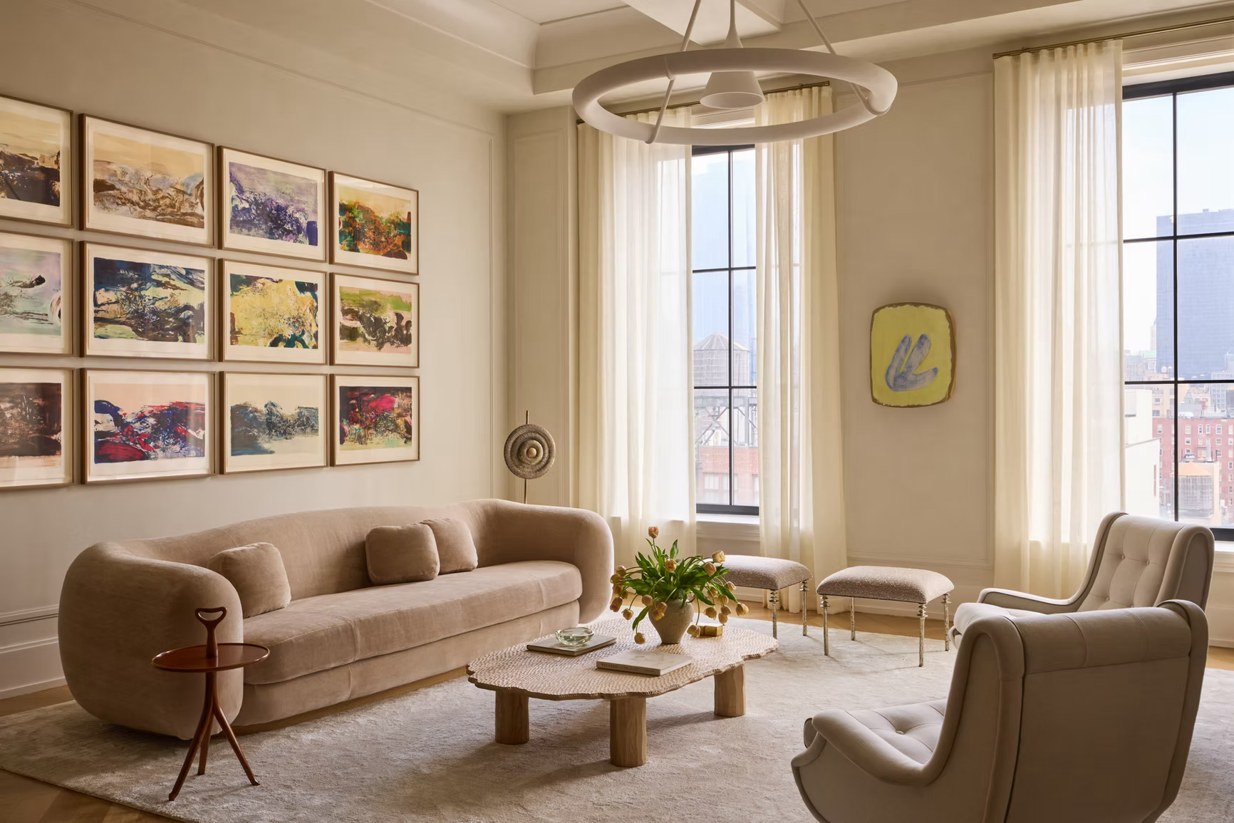 Bright transitional living room with curved beige sofa, neutral palette, abstract gallery wall art, round coffee table, and large windows with sheer curtains.