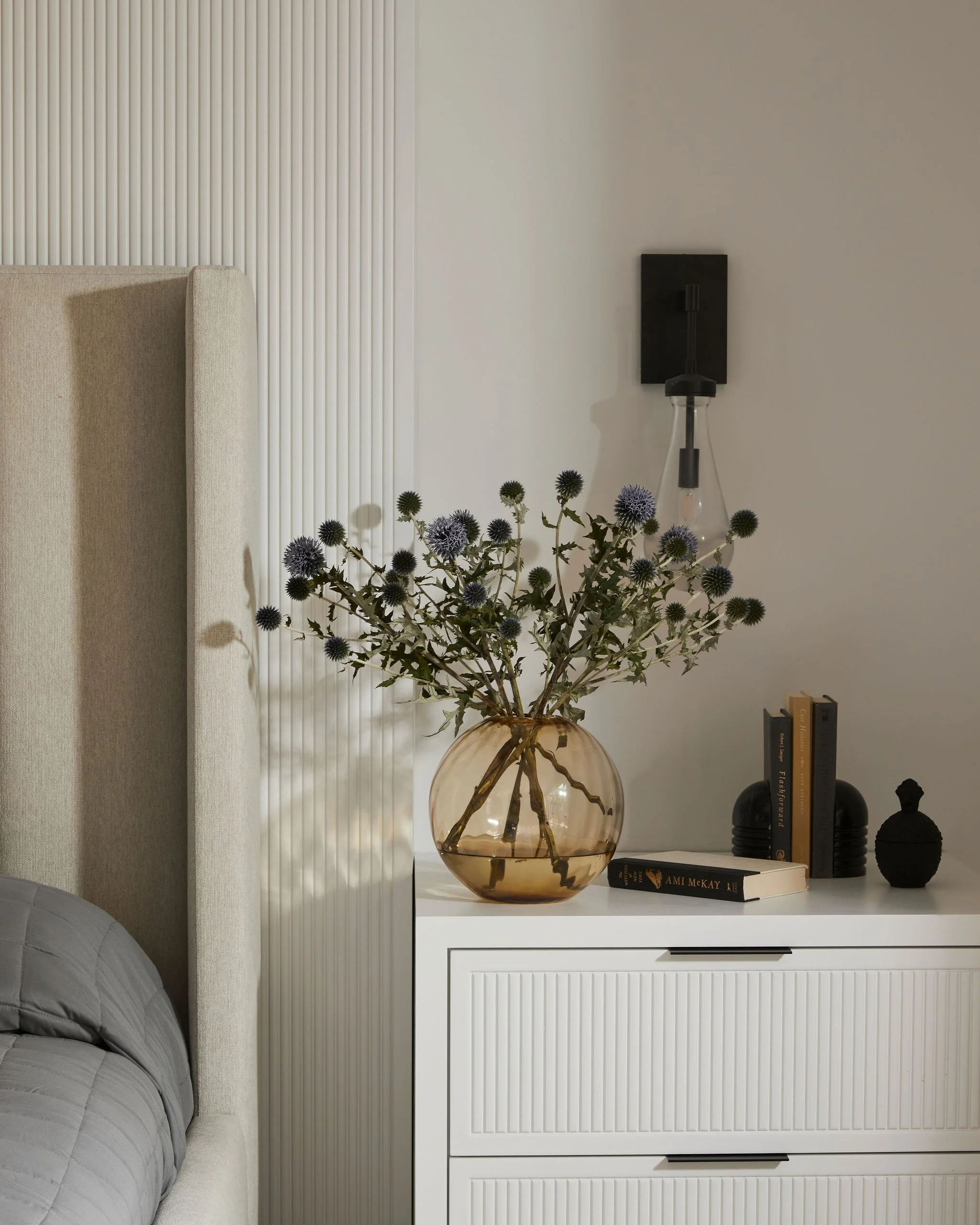 Minimal bedroom vignette with textured wall paneling, upholstered headboard, soft lighting, and styled nightstand creating a calm and layered interior.