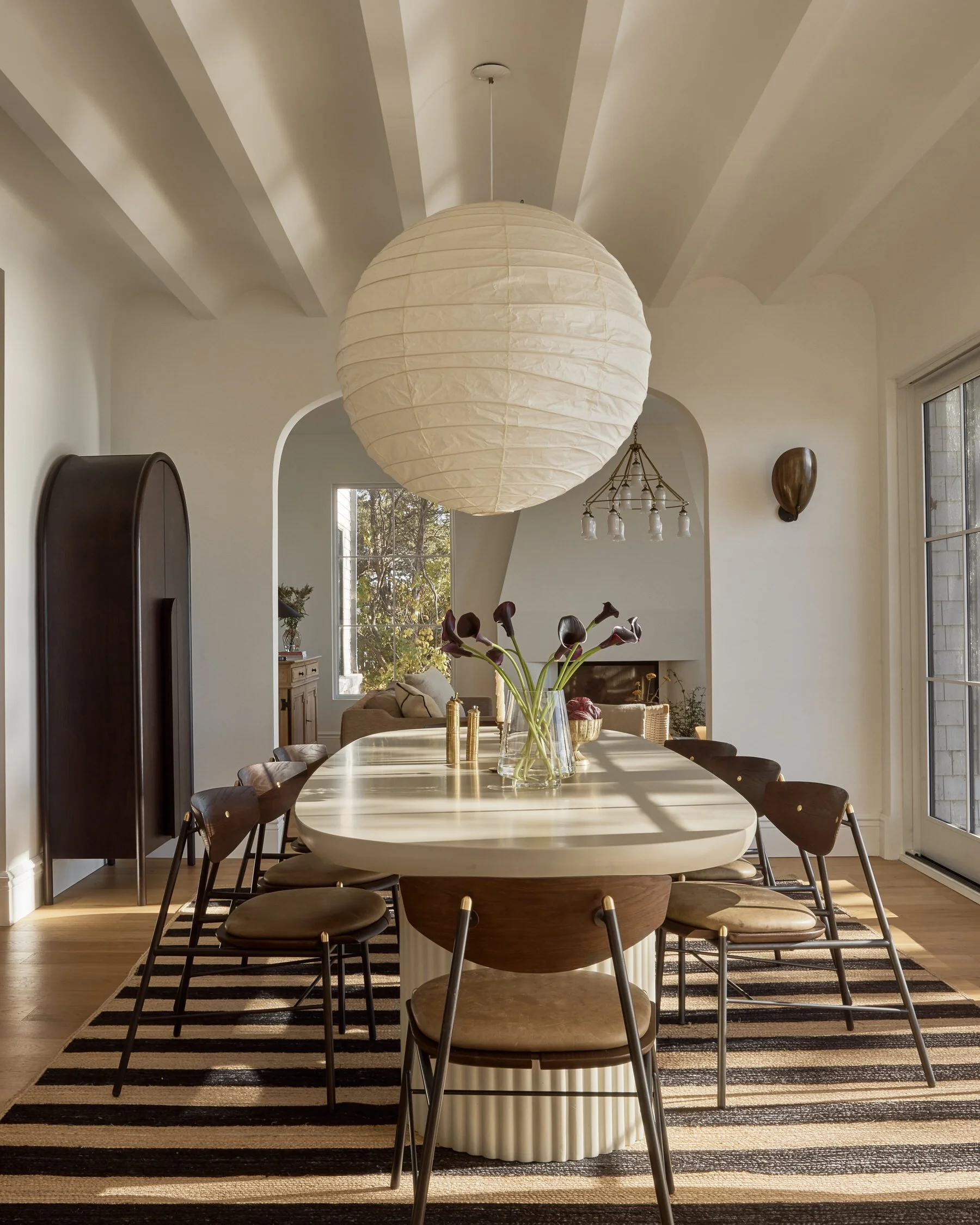 Light-filled transitional dining room featuring an oval dining table, sculptural pendant lighting, warm wood chairs, and soft neutral finishes that create a collected, cohesive space for couples with blended styles.