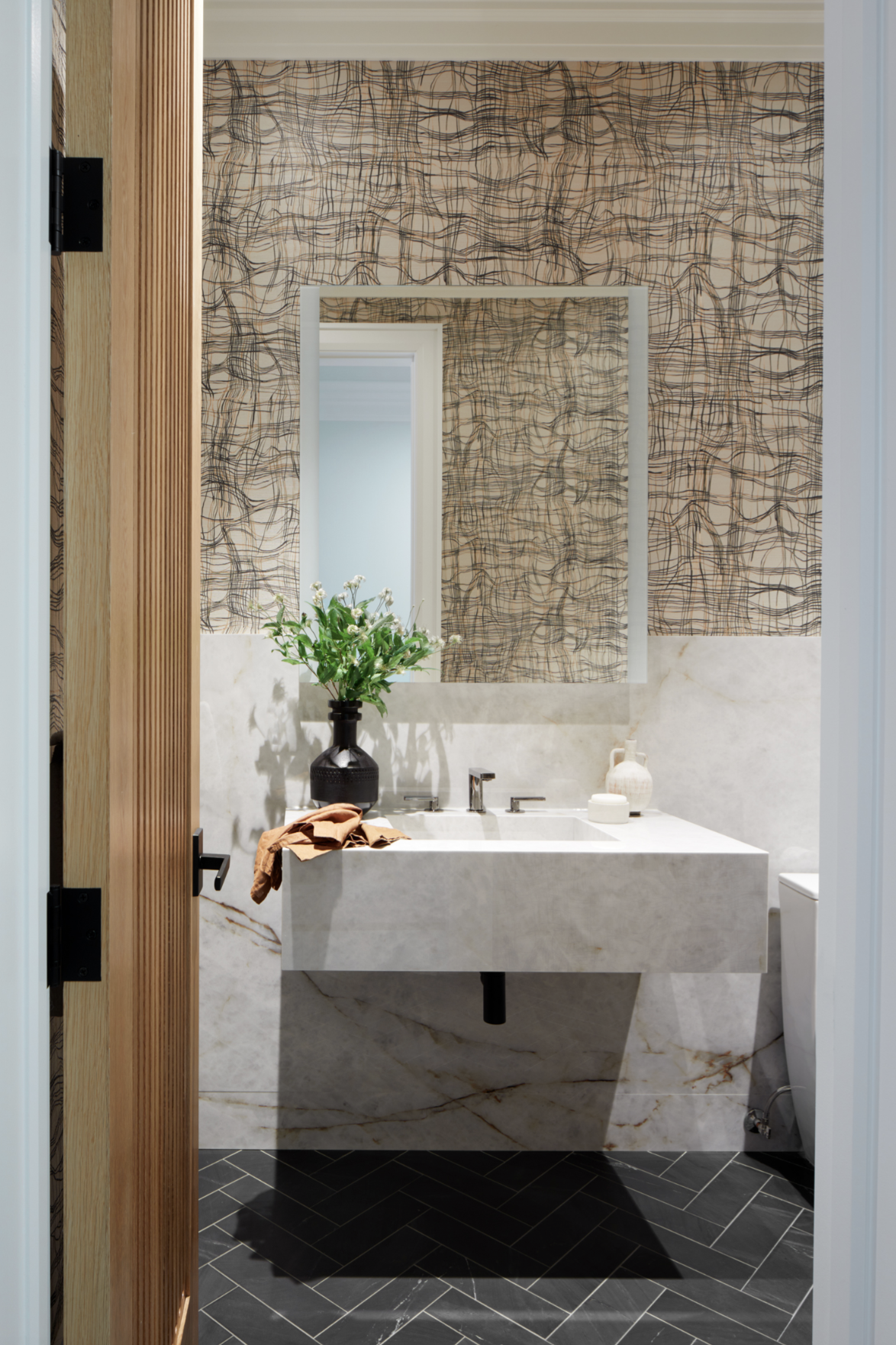 Modern powder room with marble sink, graphic wallpaper, black fixtures, and layered materials blending contemporary and classic design elements.
