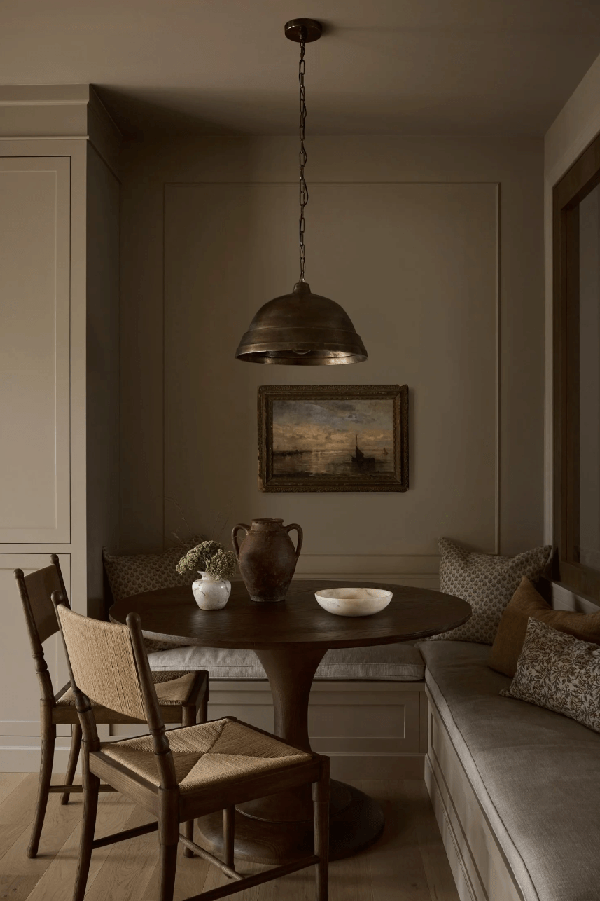 Cozy traditional breakfast nook featuring upholstered banquette seating, a round dark wood table, woven chairs, and an antique brass pendant light.