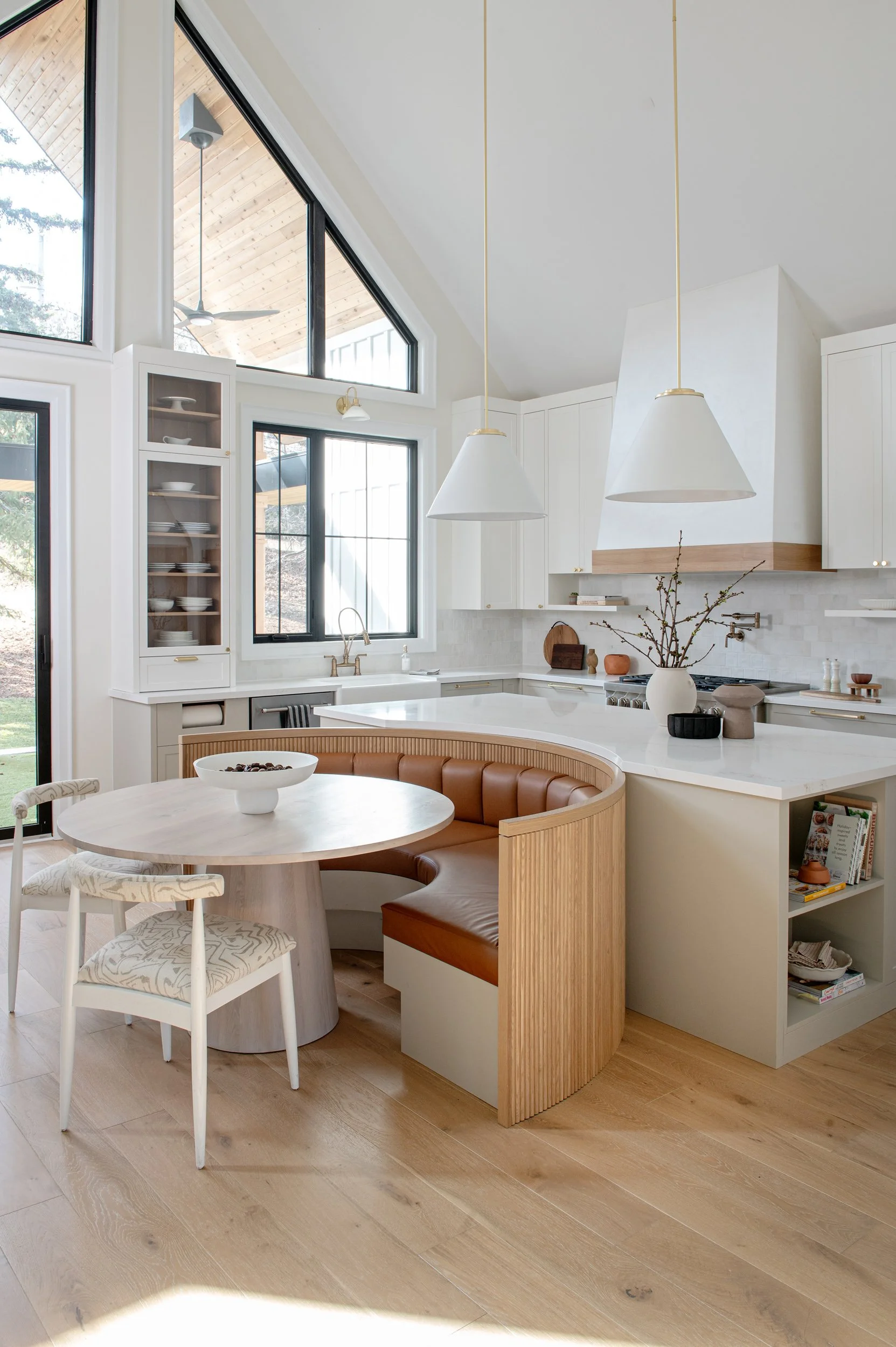 Organic modern kitchen with curved wood banquette seating, white cabinetry, large windows, and natural light creating a warm and functional gathering space.