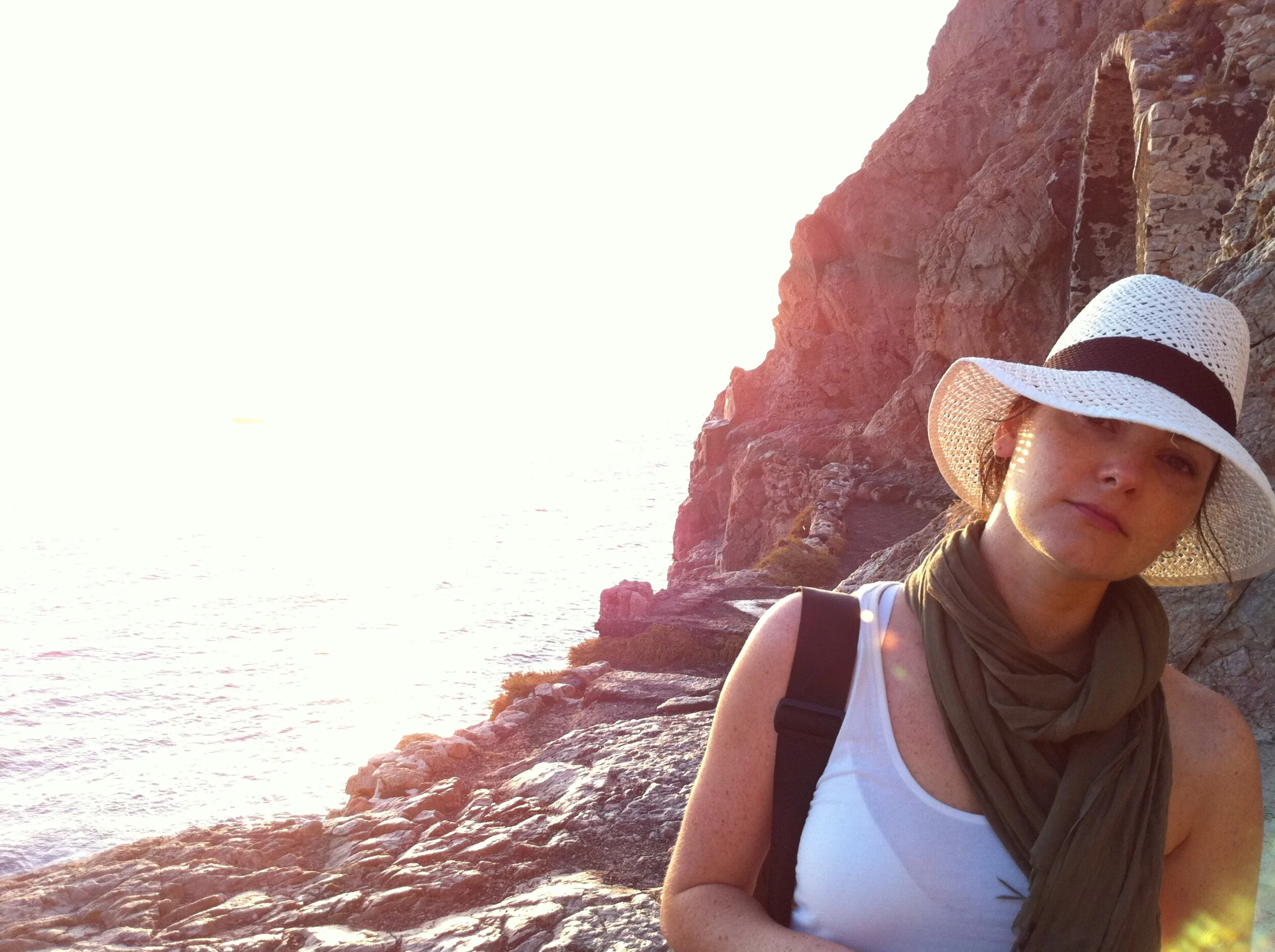 Woman wearing a wide-brimmed white hat, white tank top, and green scarf around her neck, standing against rocky cliffs near the ocean at sunset.