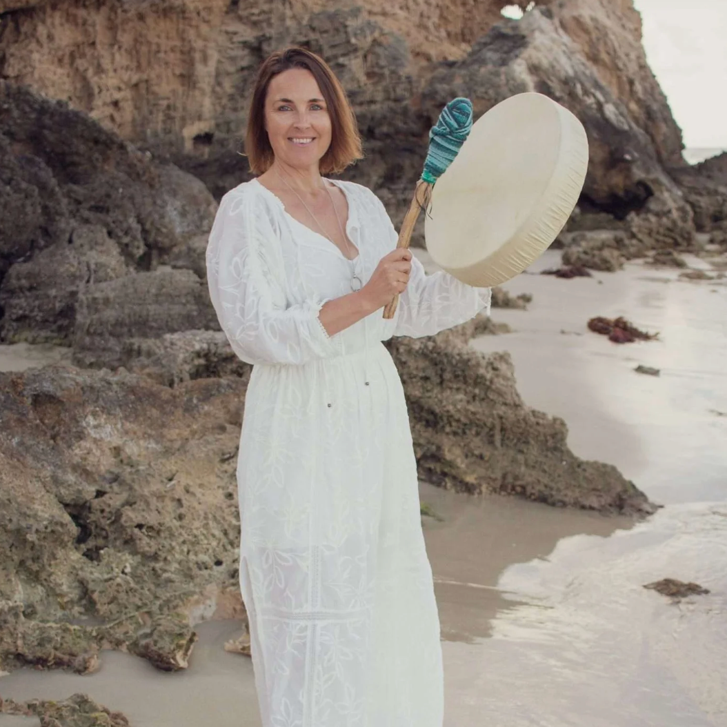Woman in a white dress holding a percussion instrument on a beach with rocks and sand.