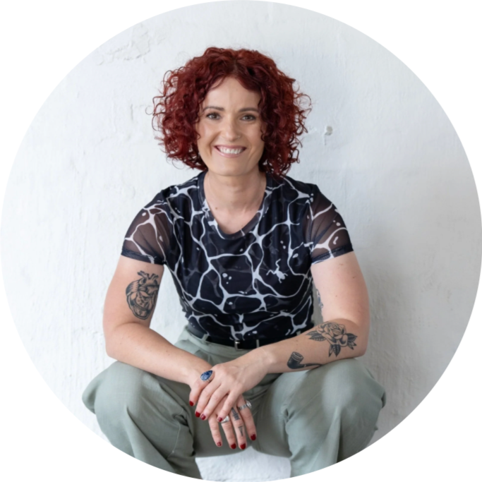 A woman with curly red hair sitting against a white wall, smiling, wearing a black and white patterned t-shirt and light-colored pants, with tattoos on her arms.