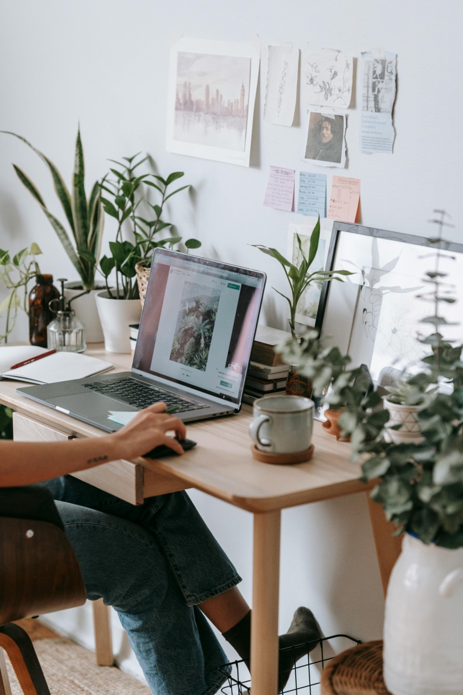 A person working at a wooden desk with a laptop, surrounded by various plants, notebooks, and decorative items, with papers and artwork pinned on a white wall behind.