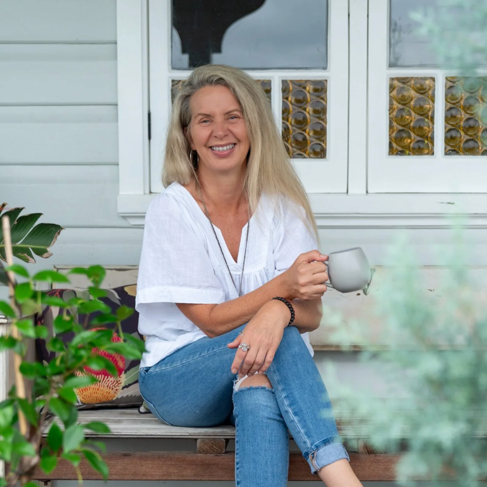 A woman with long gray hair sitting outside on a porch, smiling, holding a mug, wearing a white top and ripped jeans.