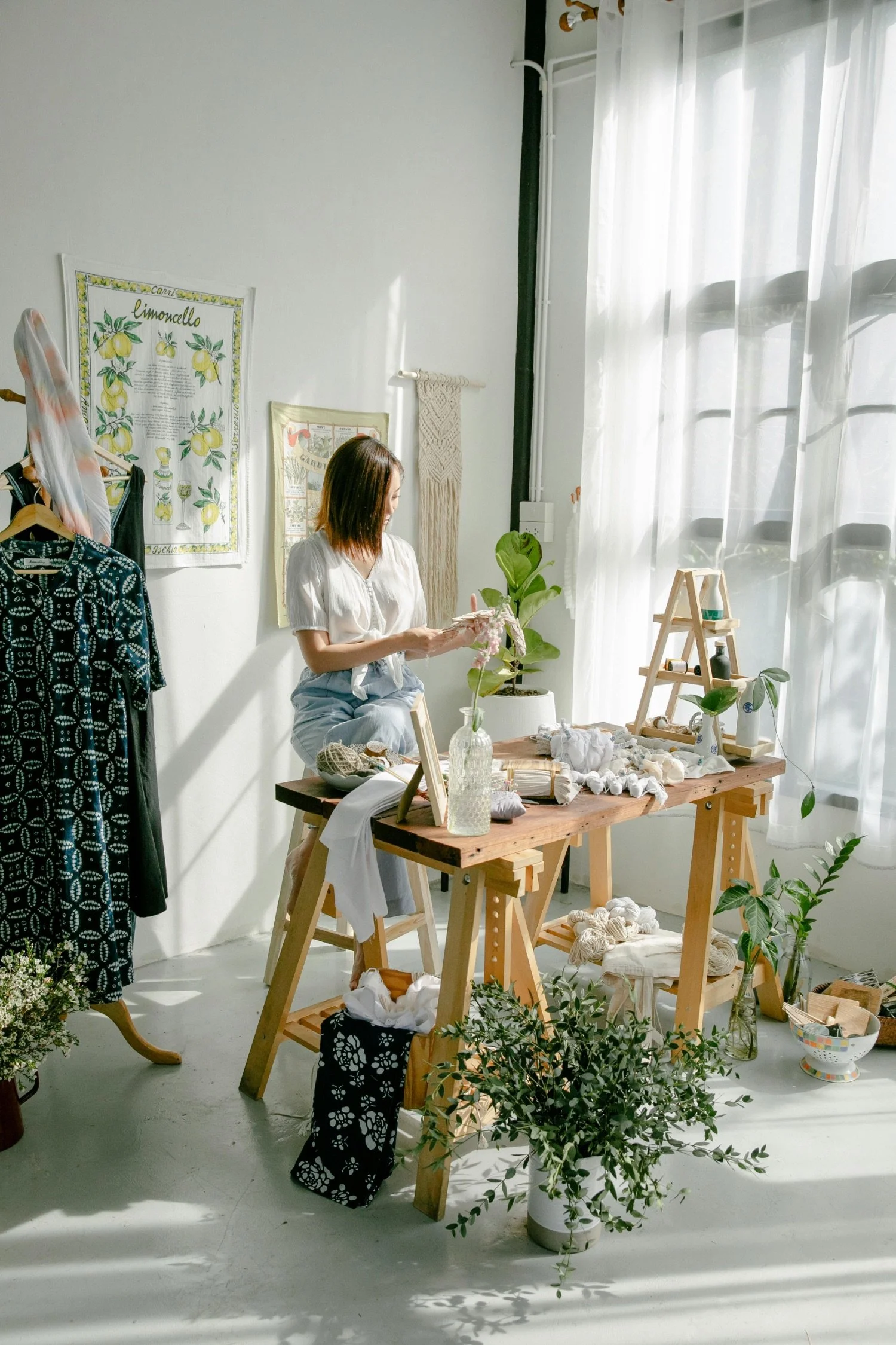 Woman sitting at a wooden table with crafting supplies, surrounded by plants, posters, and hanging garments, in a bright room with white walls and curtains.