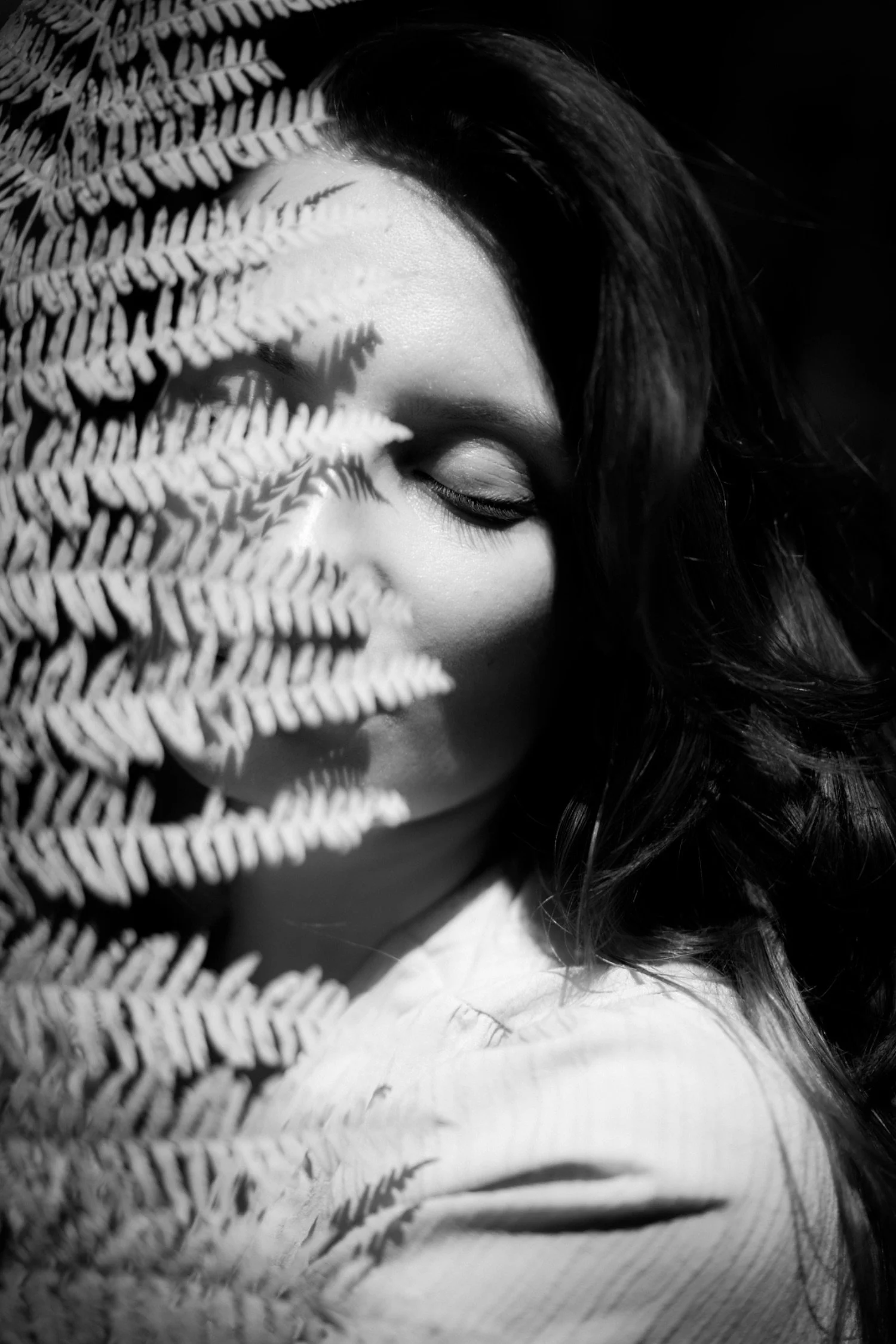A black and white photograph of a woman with closed eyes, partially covered by fern leaves.