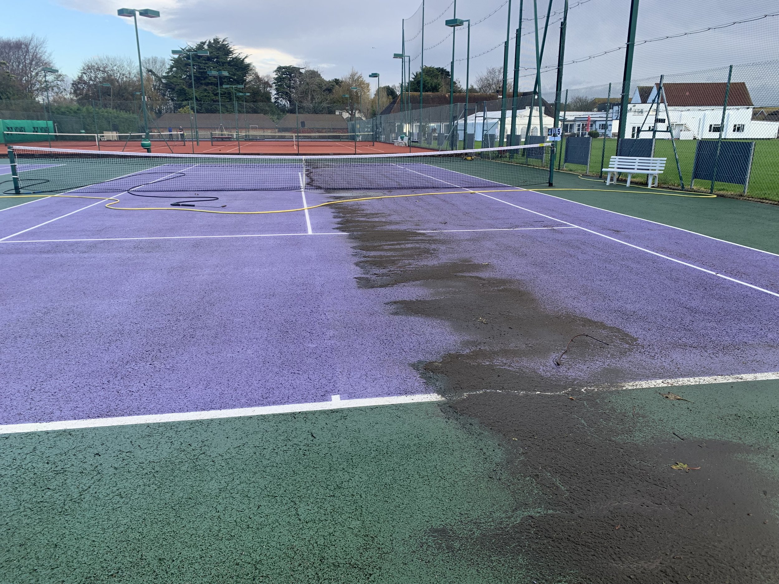 A tennis court with purple and green surface, damp in the middle, set outdoors with trees, houses, and a cloudy sky in the background.
