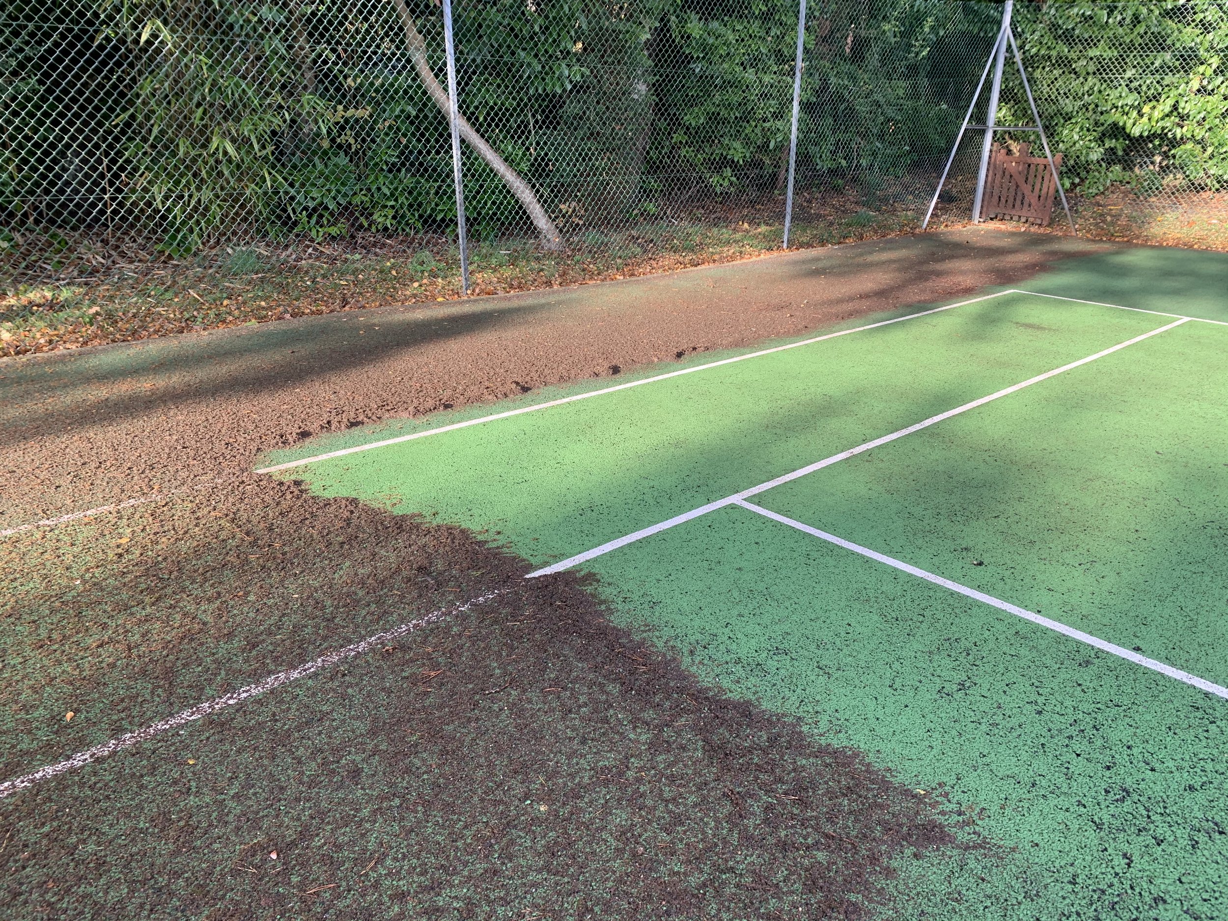 Dirt and worn green tennis court with white lines, surrounded by a chain-link fence and lush greenery.