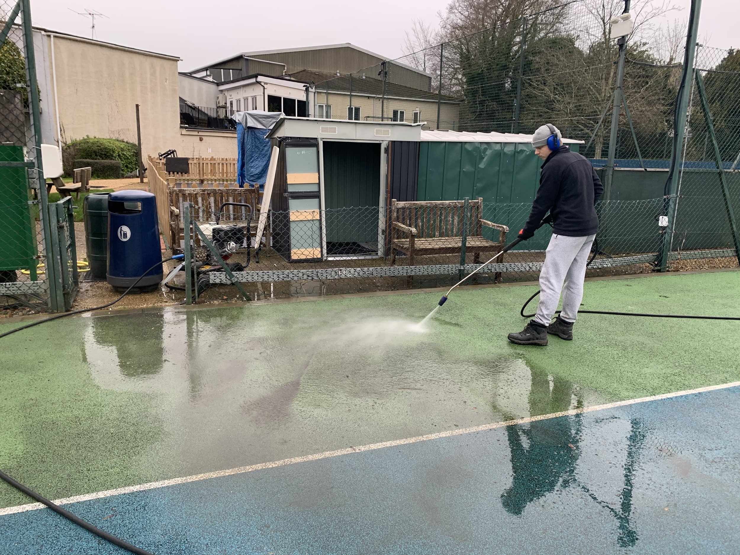 A person pressure washing a sports court with a green and blue surface, wearing gray sweatpants, black boots, a black hoodie, gray beanie, and blue ear protection.