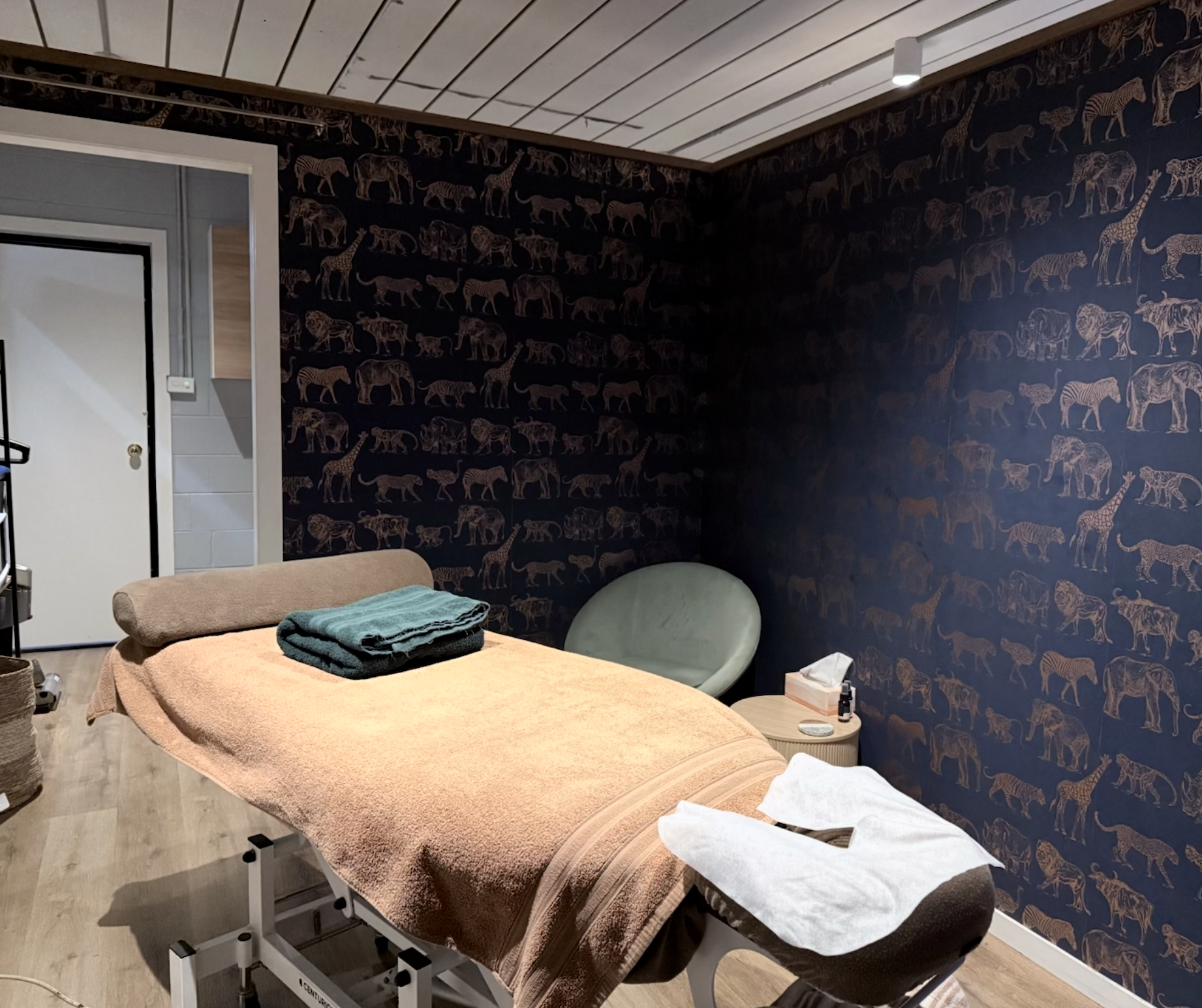 Core Bodywork treatment room featuring a massage table, folded towels, seating and wallpaper.