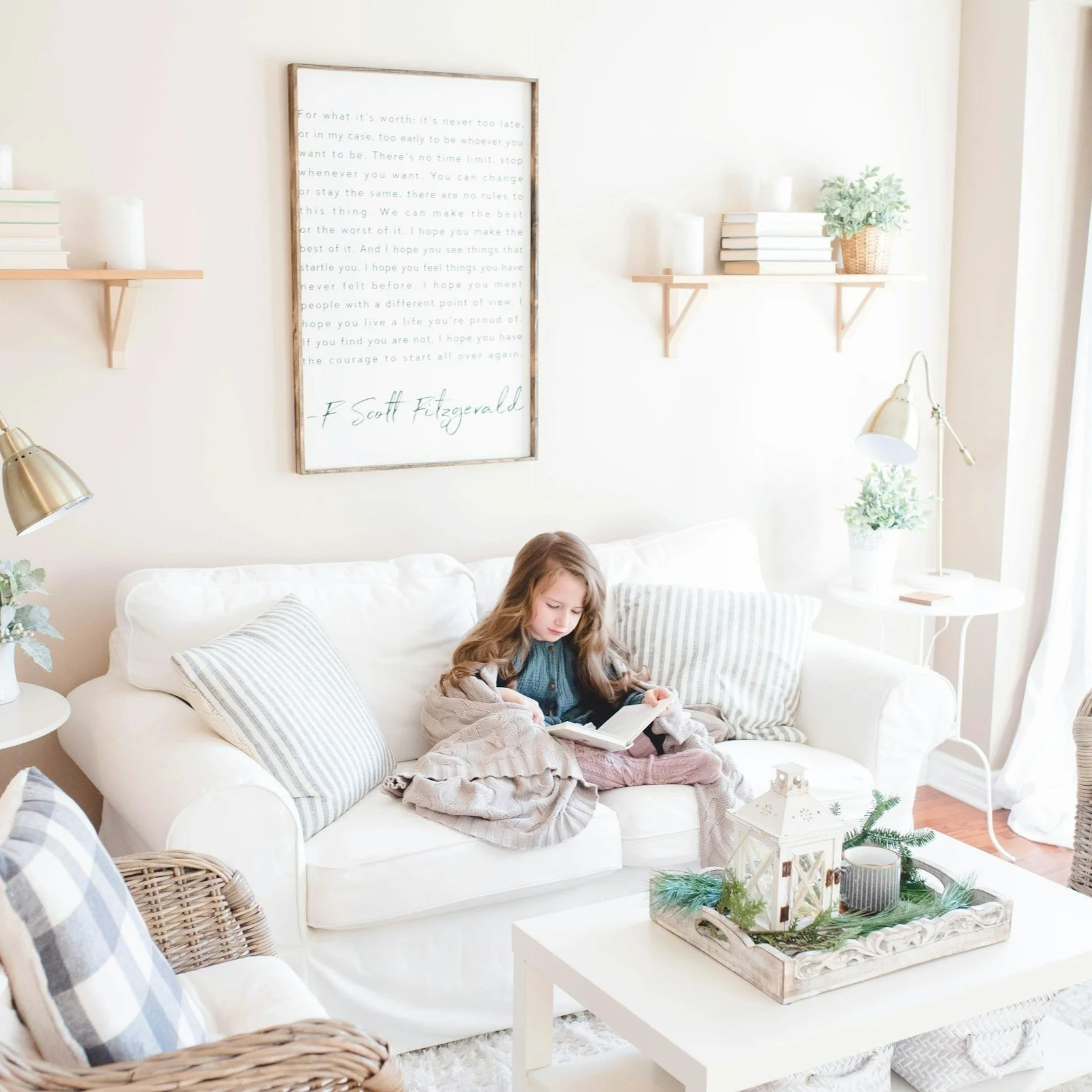 A young girl sitting on a white sofa reading a book in a bright, cozy living room decorated in a neutral, farmhouse style with plush cushions, wall shelves with books and plants, and a coffee table.