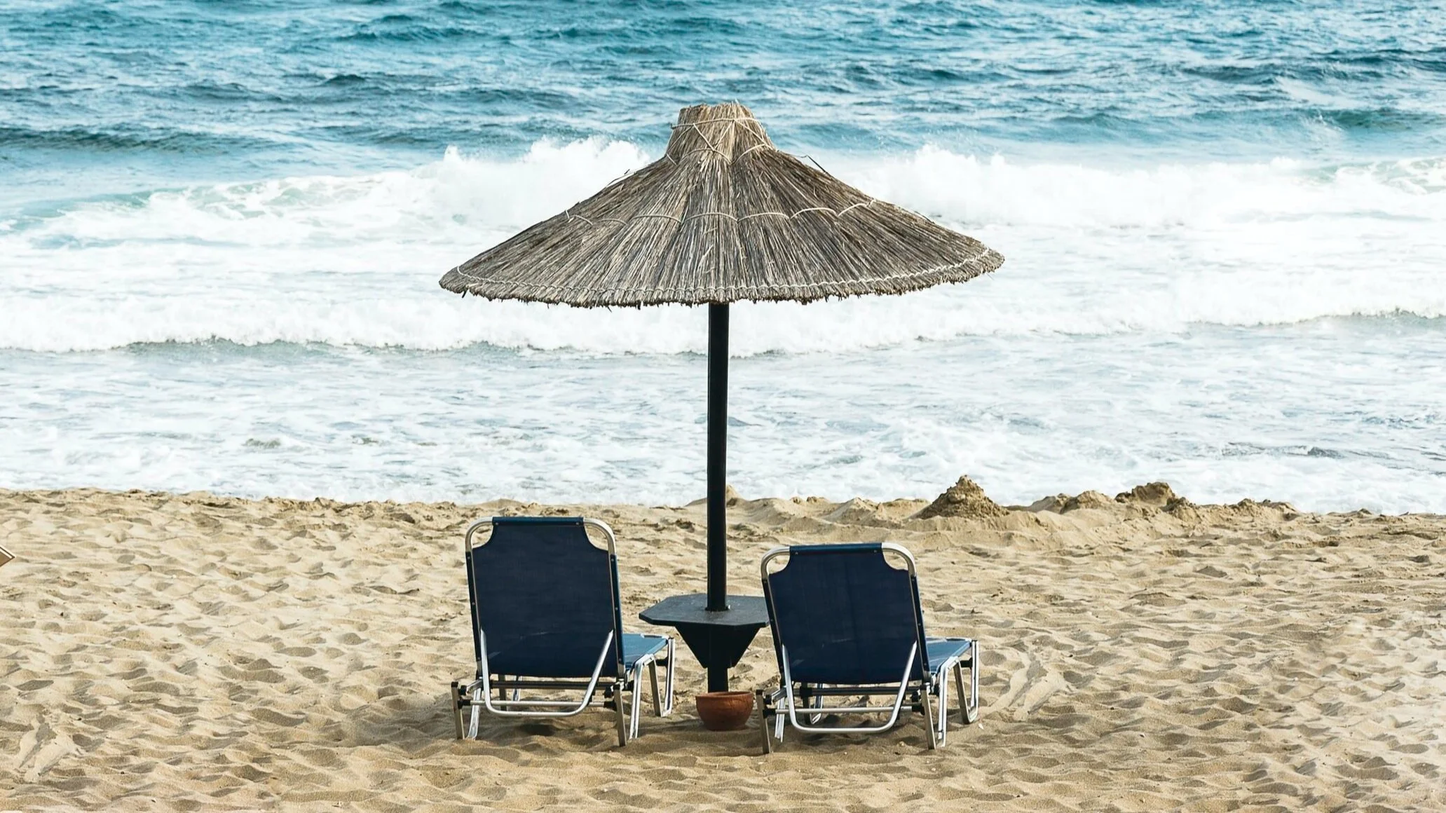 Sandstrand ved havet med to tomme solstolar og ein parasoll.  Foto: @kmile_ch / Unsplash