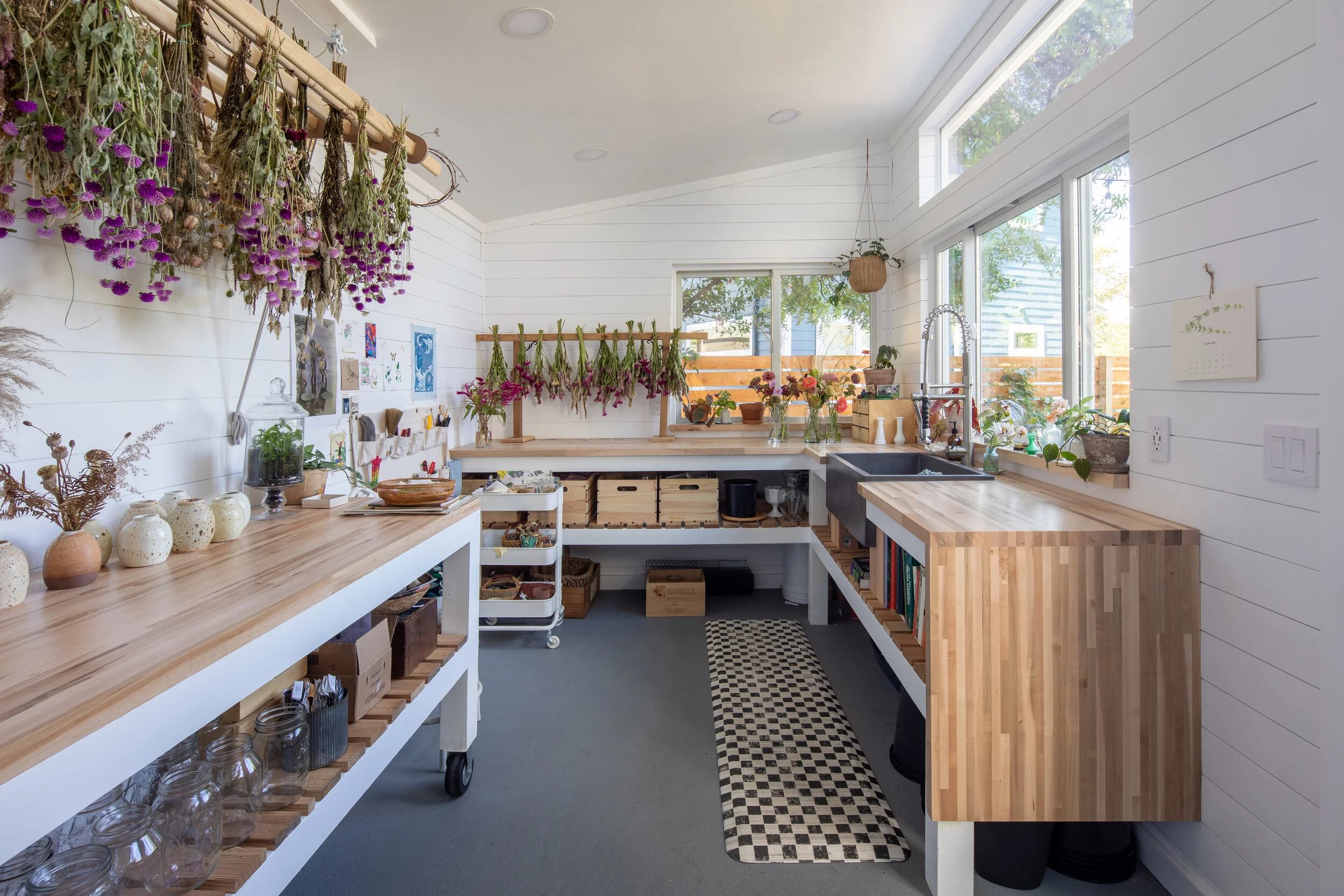 Interior of a bright, organized botanical workshop with wooden tables, drying herbs and flowers hanging, potted plants, and storage baskets.